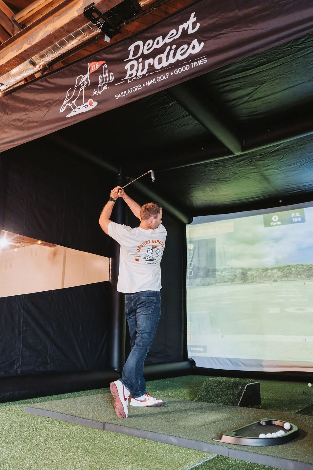 A man playing golf using an indoor golf simulator, wearing a white t-shirt with the 'Desert Birdies' logo, and standing on artificial turf with golf balls and a tracking device on the floor.
