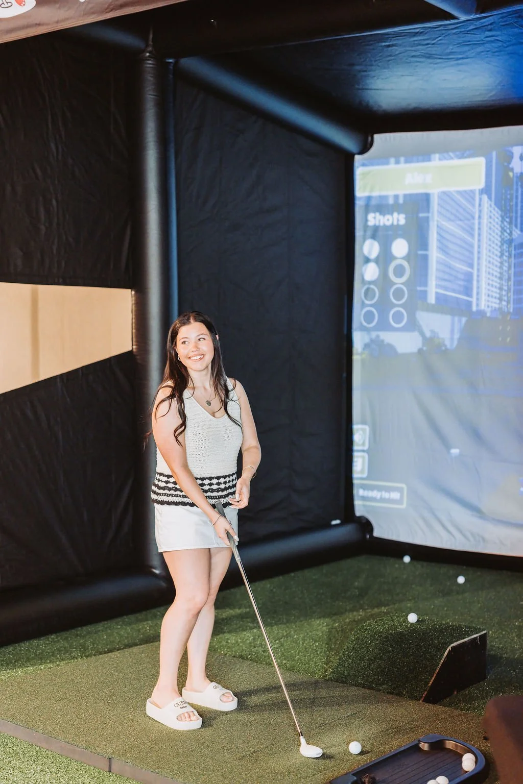 A woman with long dark hair, smiling, standing on an indoor golf simulator mat, holding a golf club, surrounded by golf balls, with a large screen displaying a virtual golf course in the background.