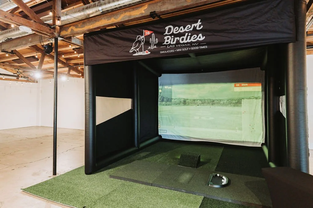 Indoor golf simulator setup with a large screen displaying a golf course, surrounded by black inflatable walls, and a sign above reading 'Desert Birdies LAS VEGAS, NV'.