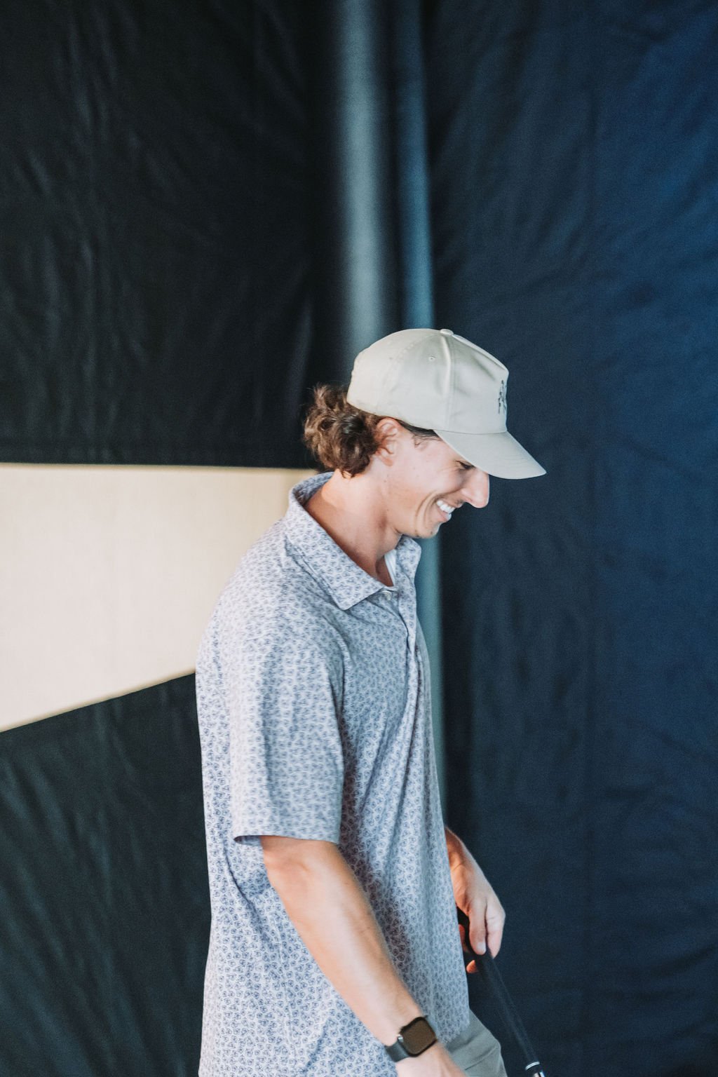 Young man smiling, wearing a baseball cap and patterned shirt, standing against a dark background.