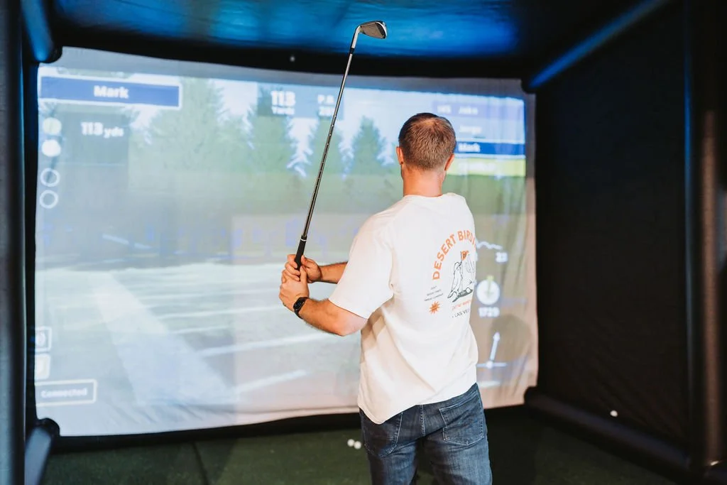 Man practicing golf with a simulator in an indoor setup, wearing a white T-shirt and jeans, holding a golf club and facing a large digital screen.