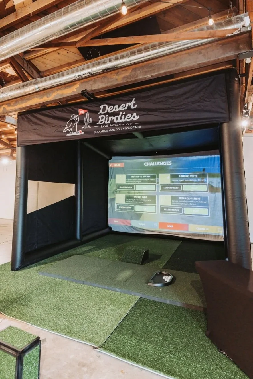 Indoor golf simulator setup with a black inflatable enclosure, artificial turf, and a large screen displaying golf challenges at Desert Birdies in Las Vegas, NV.