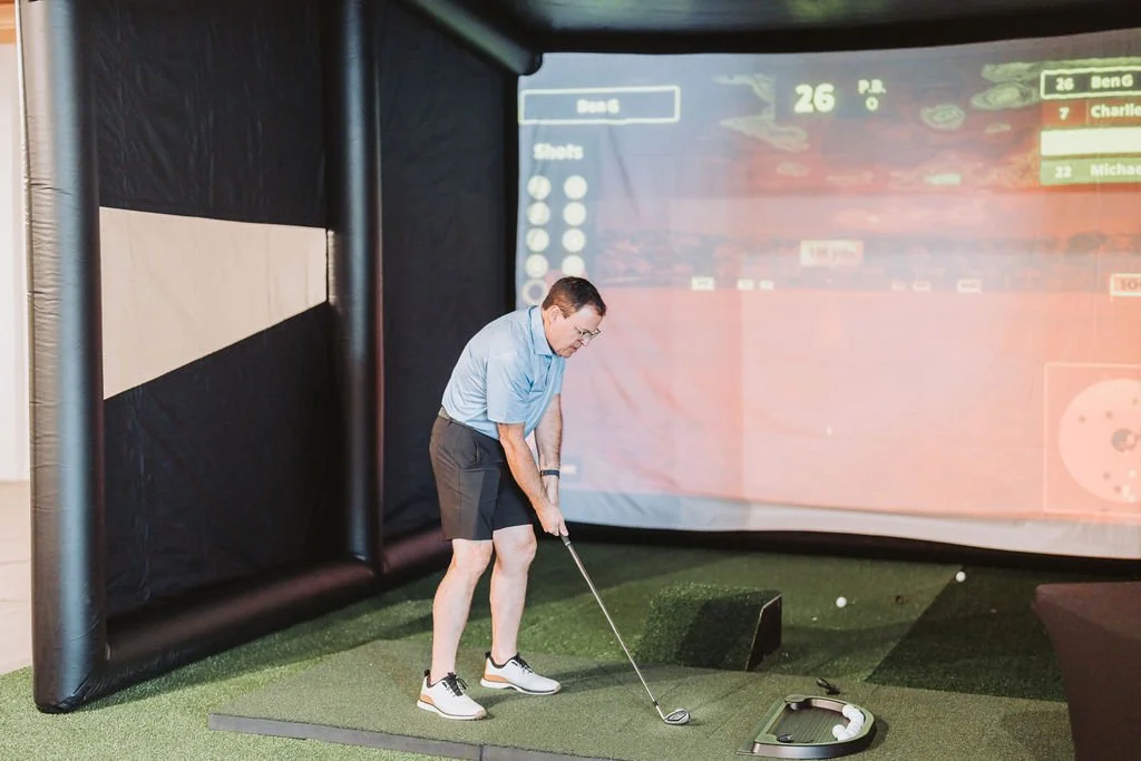 Man practicing golf swing indoors on artificial grass, with golf balls and a screen displaying a golf game in the background.