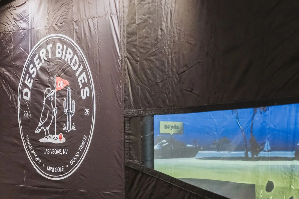 Part of an indoor golf simulator with a black wall displaying a logo for Desert Birdies Golf Society, featuring a bird, cactus, and golf ball, along with the text 'Las Vegas, NV 2026' and 'Simulators - Mini Golf - Good Times'.