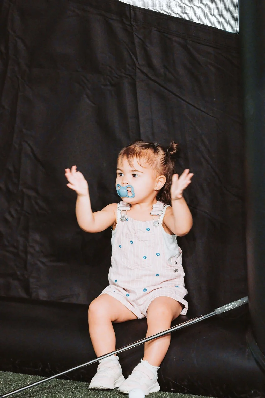 A young girl sitting with her hands raised in front of a black backdrop, wearing a white dress with blue flowers, a pacifier in her mouth, and holding a golf club, with a golf ball on the ground.