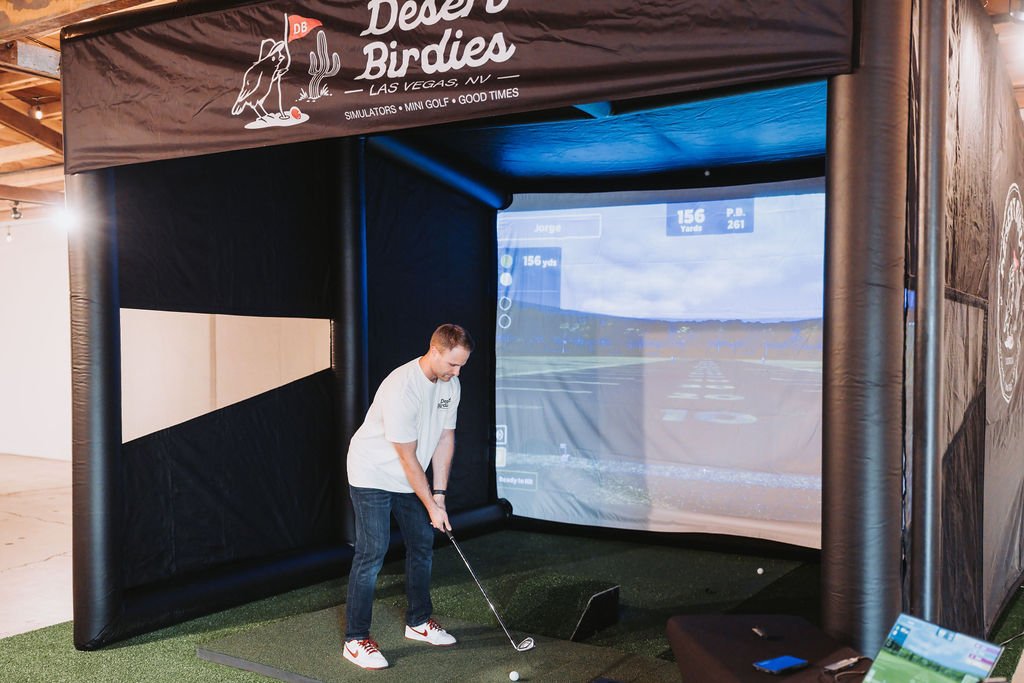A young man practicing golf swings at an indoor simulator called Desert Birdies with a large screen showing a virtual golf course.