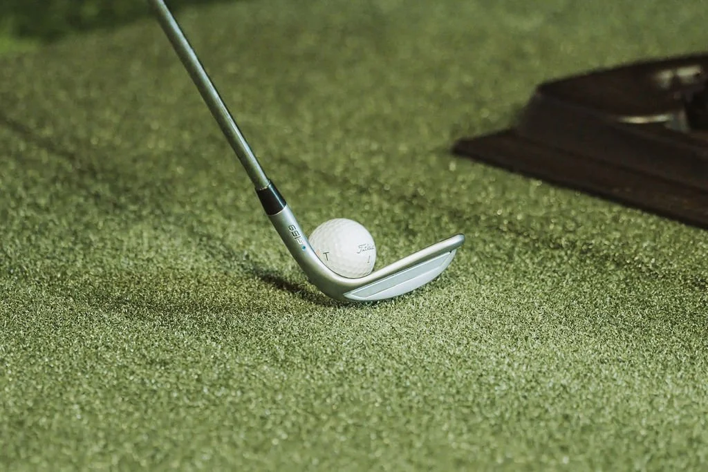 Close-up of a golf club and golf ball on a putting green with a brown golf tee box in the background.