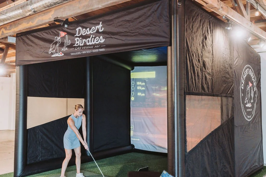 An indoor golf simulator setup called 'Desert Birdies' with a woman preparing to hit a golf ball inside a tented enclosure with artificial grass and a large screen display.