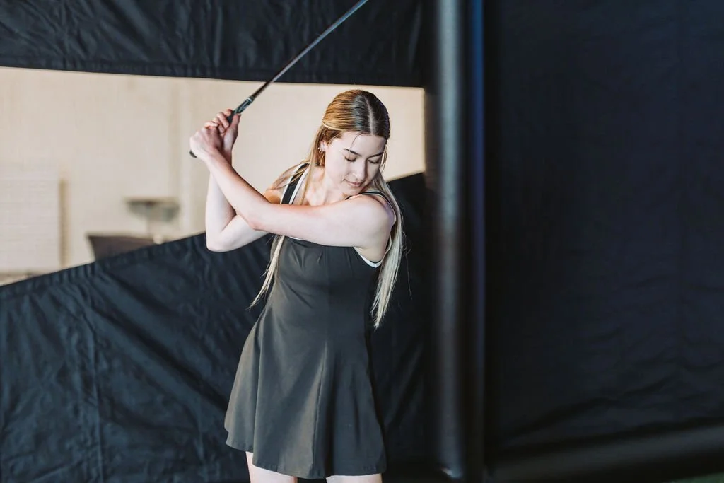 A woman in a black athletic dress practicing golf swing indoors with a black curtain background.