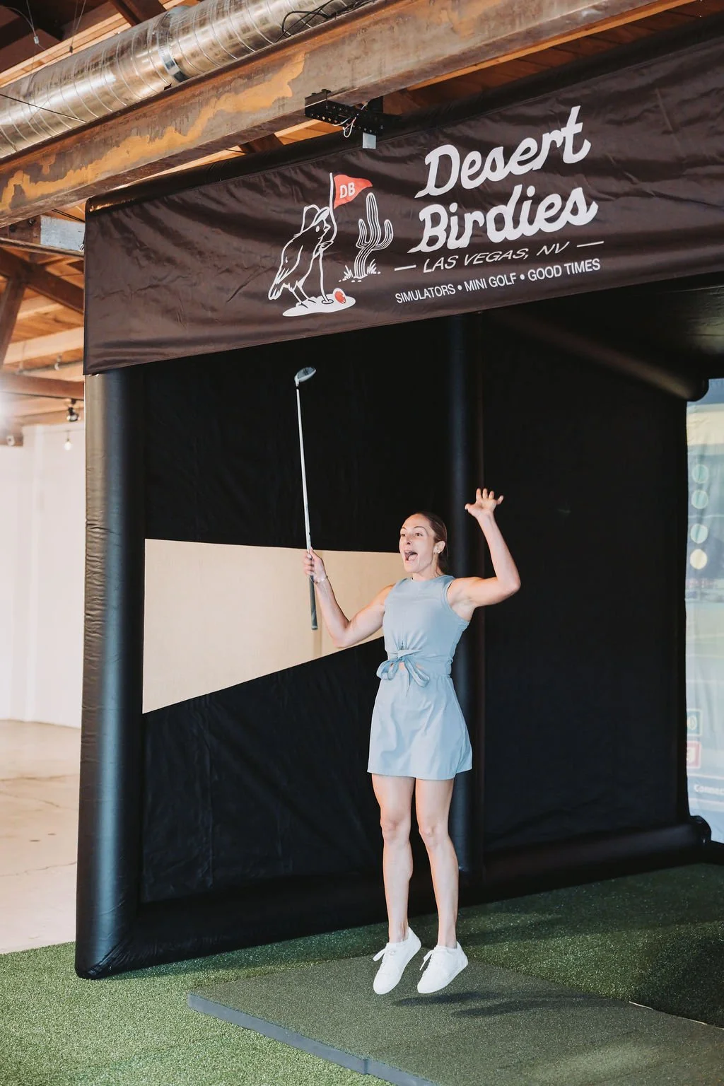 A woman jumping in front of an indoor golf simulator at Desert Birdies in Las Vegas, NV, holding a golf club and appearing excited.