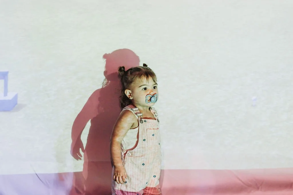 A young girl with a pacifier in her mouth standing against a plain, light-colored wall, casting a shadow behind her.