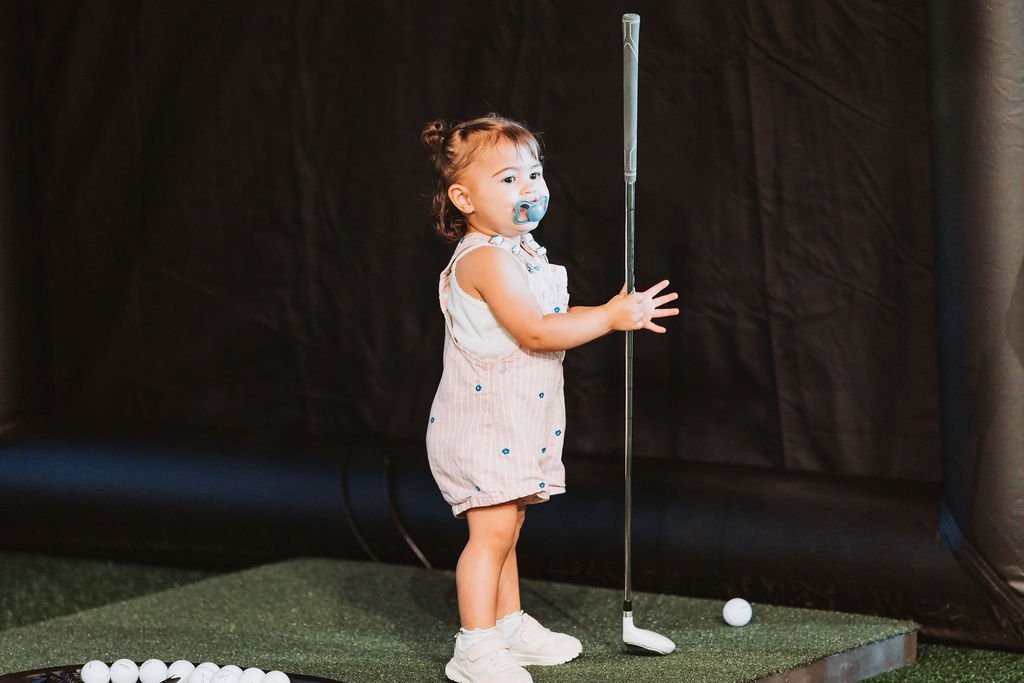 A young girl with a pacifier holding a golf club on a miniature golf course, looking to the side, with golf balls in front of her.