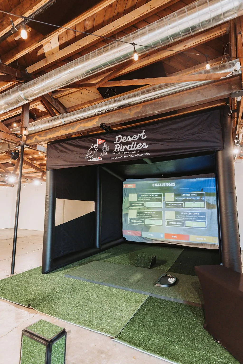 Indoor golf simulation game station with a screen displaying challenges, turf mat, and banner reading 'Desert Birdies, Las Vegas, NV, Simulators, Mini Golf, Good Times.'