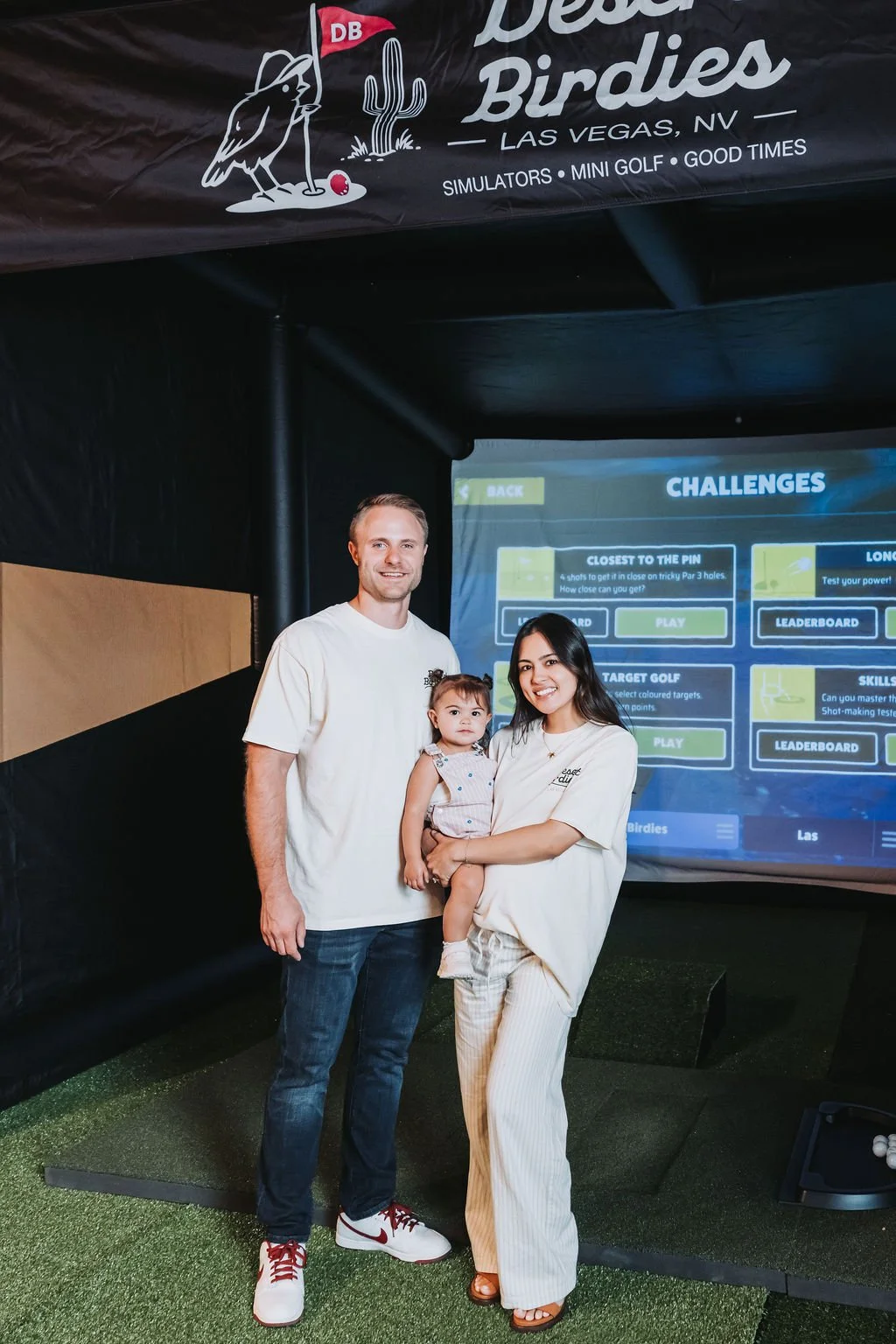 A family of three standing in front of an arcade game screen, at Desert Birdies mini golf in Las Vegas, NV. The man is wearing a white t-shirt and jeans, the woman is wearing a light-colored top and striped trousers, and they are holding a young girl in a light-colored dress. In the background, there is a banner with the logo of Desert Birdies featuring a bird and a cactus.
