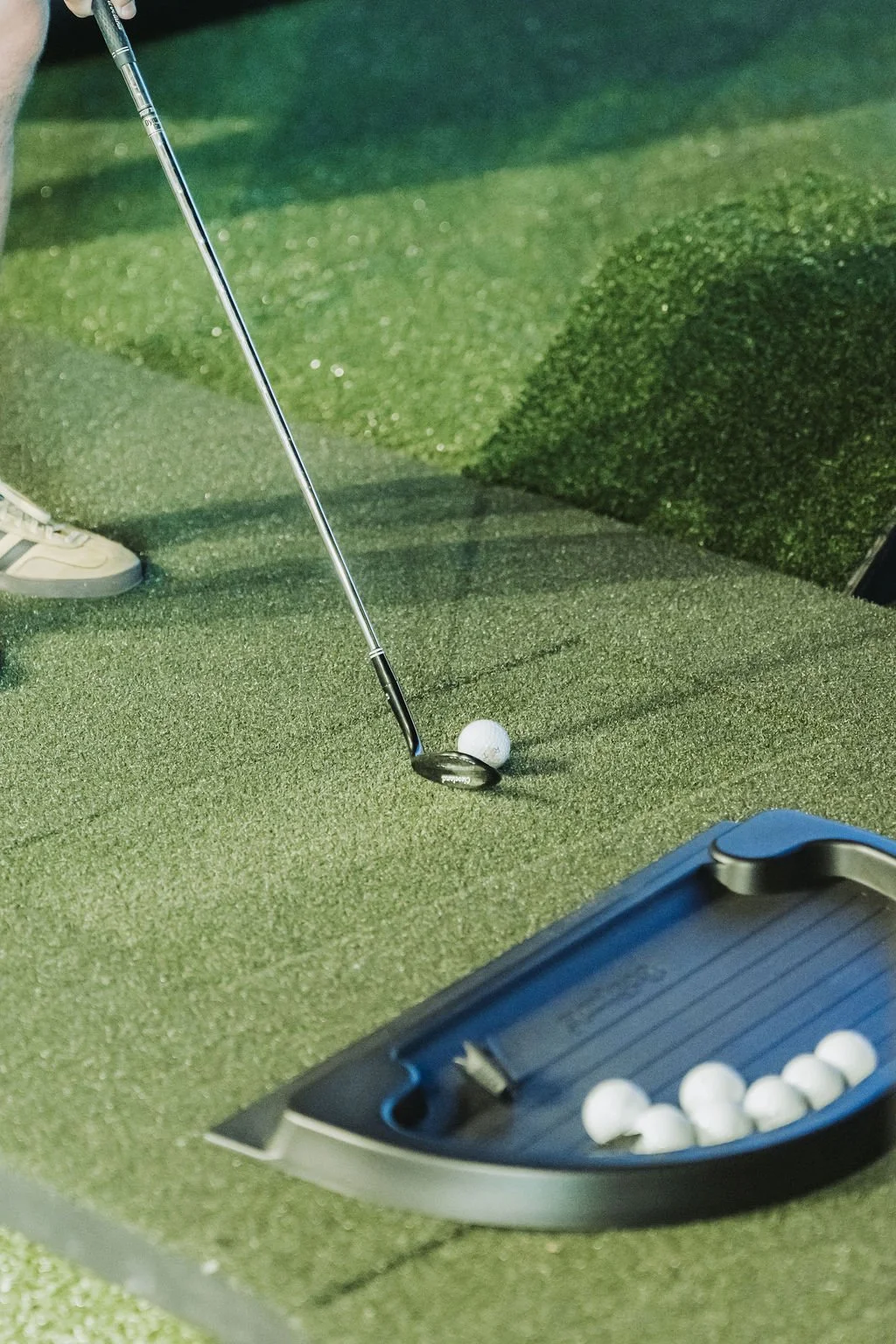A golf club poised to hit a golf ball on an artificial green with a tray of additional golf balls nearby.