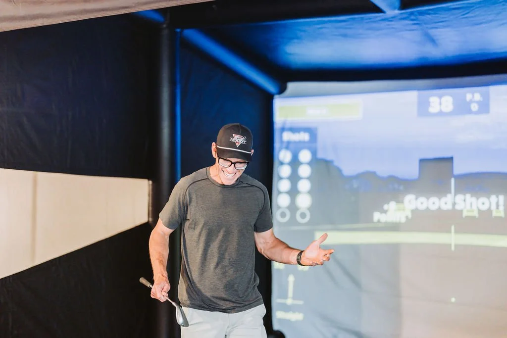 A young man in a baseball cap and glasses smiling inside a golf simulation room.