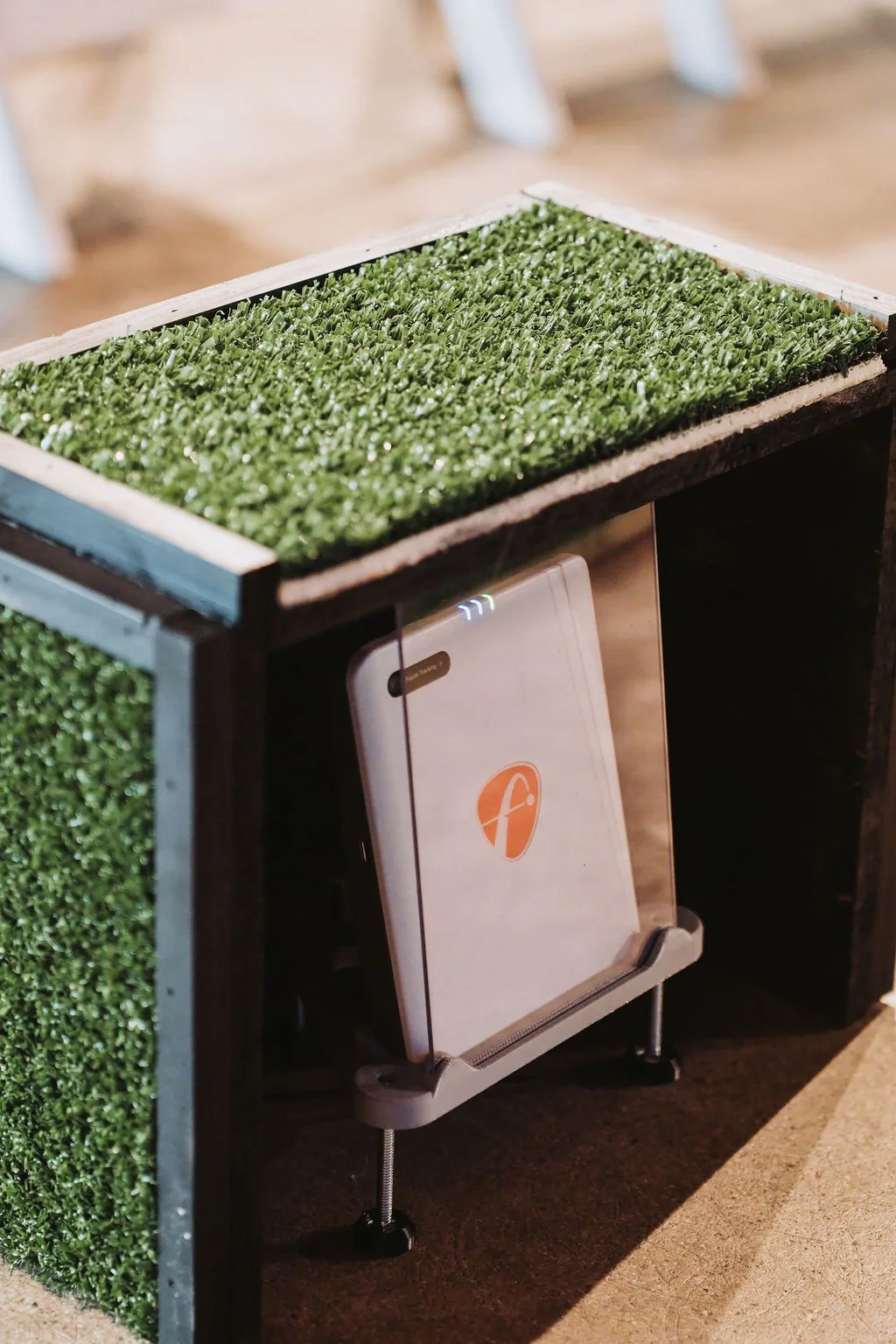A tablet with a white cover and a logo, placed on a stand beneath a table with fake grass top.