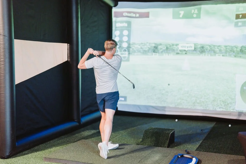 A man playing virtual golf indoors at an indoor golf simulation center, wearing casual athletic clothes, swinging a golf club to hit a ball towards a projected screen showing a virtual golf course.