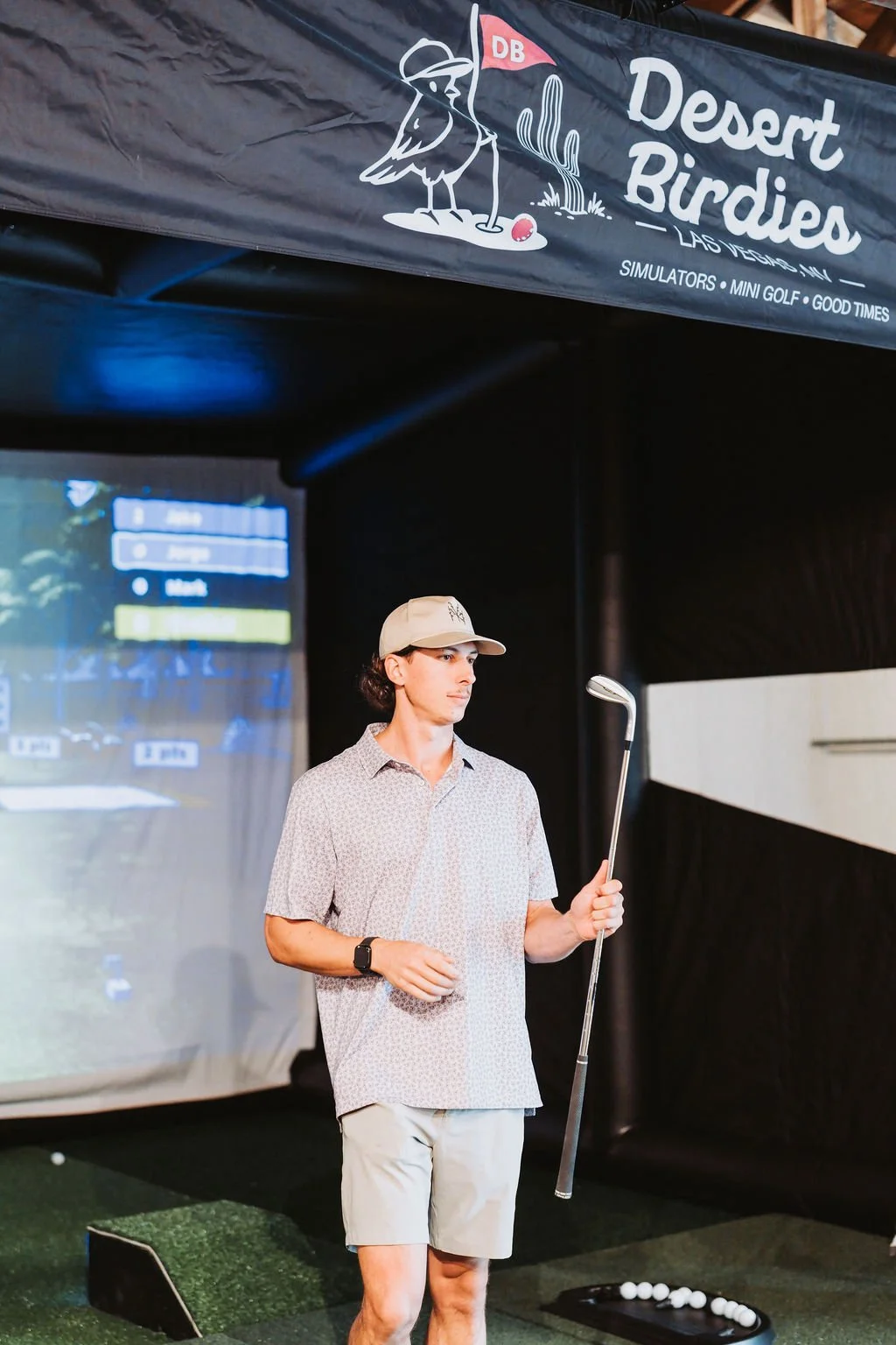 A man in a beige cap, patterned shirt, and shorts holding a golf club at an indoor golf simulator. There is a golf ball tray on the ground nearby and a large screen in the background showing a golf course.