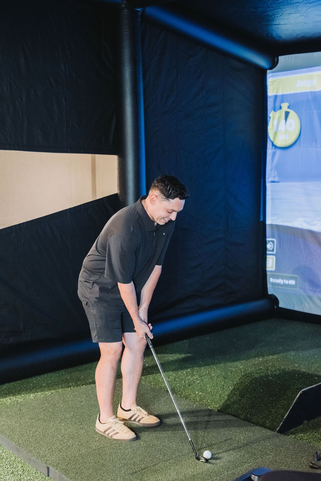 A young man in a black polo shirt and shorts is playing indoor golf on a synthetic green turf, preparing to take a shot with a golf club.