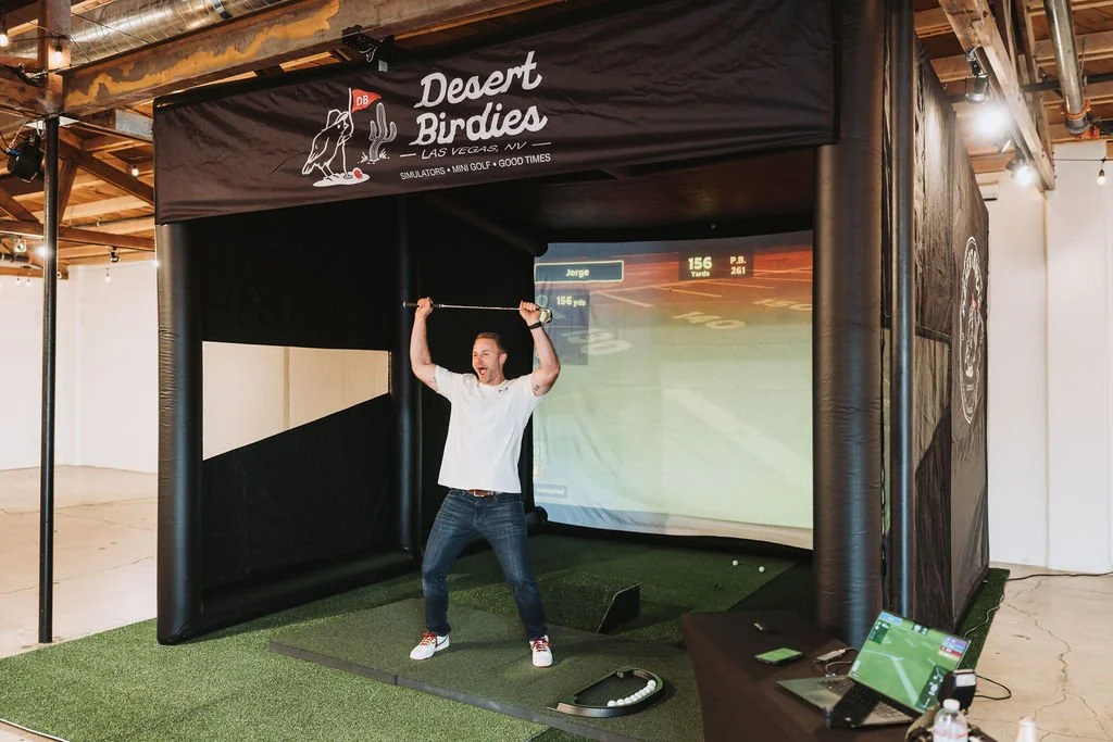 A man in a white shirt and jeans playing indoor golf at a simulator in a room with string lights, wooden beams, and a mock golf course projected on the screen.