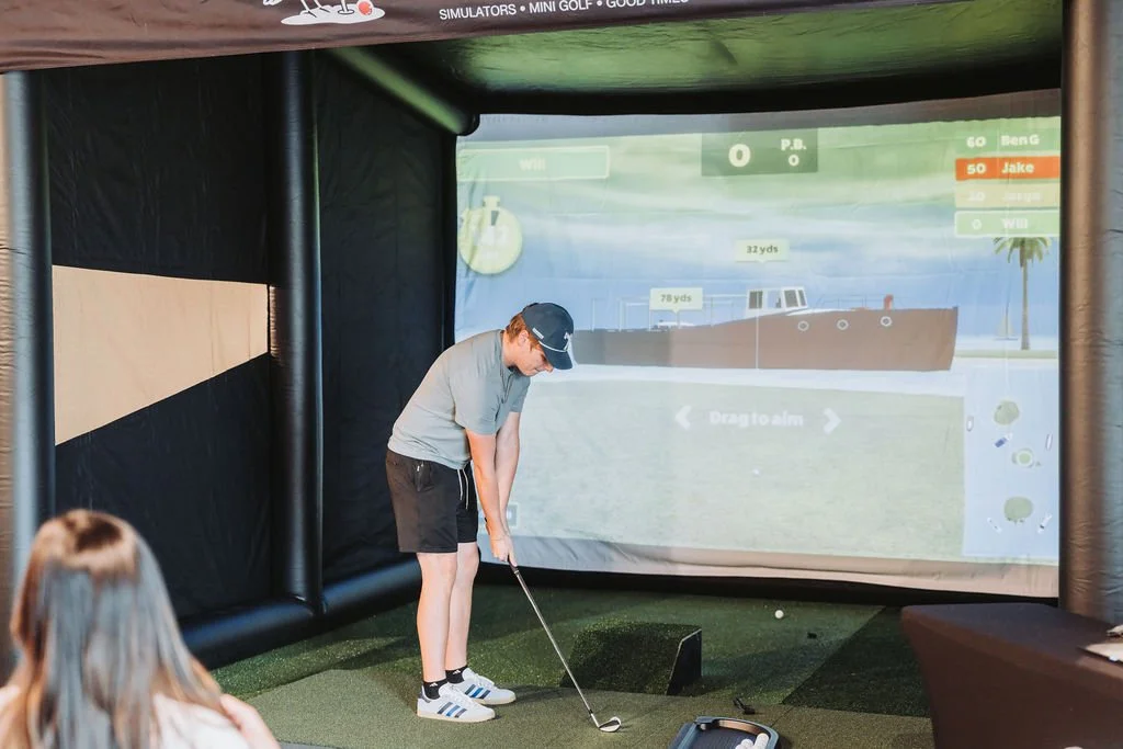 A young man wearing a baseball cap, gray shirt, black shorts, and golf shoes is playing an interactive golf simulation game indoors. He is preparing to hit a golf ball on a virtual golf course projected on the wall, with a person seated in the foregr