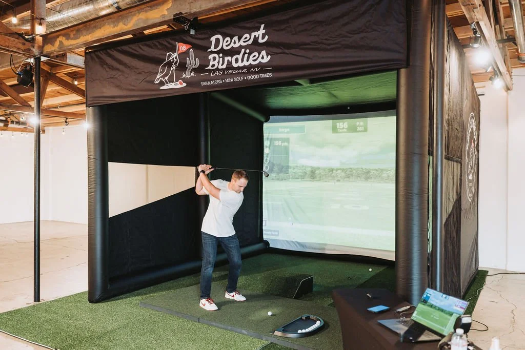 An indoor golf simulator setup with a person practicing a golf swing inside a black enclosure labeled Desert Birdies, Las Vegas. The setup includes a large screen displaying a golf course, artificial green turf, and golf training equipment.