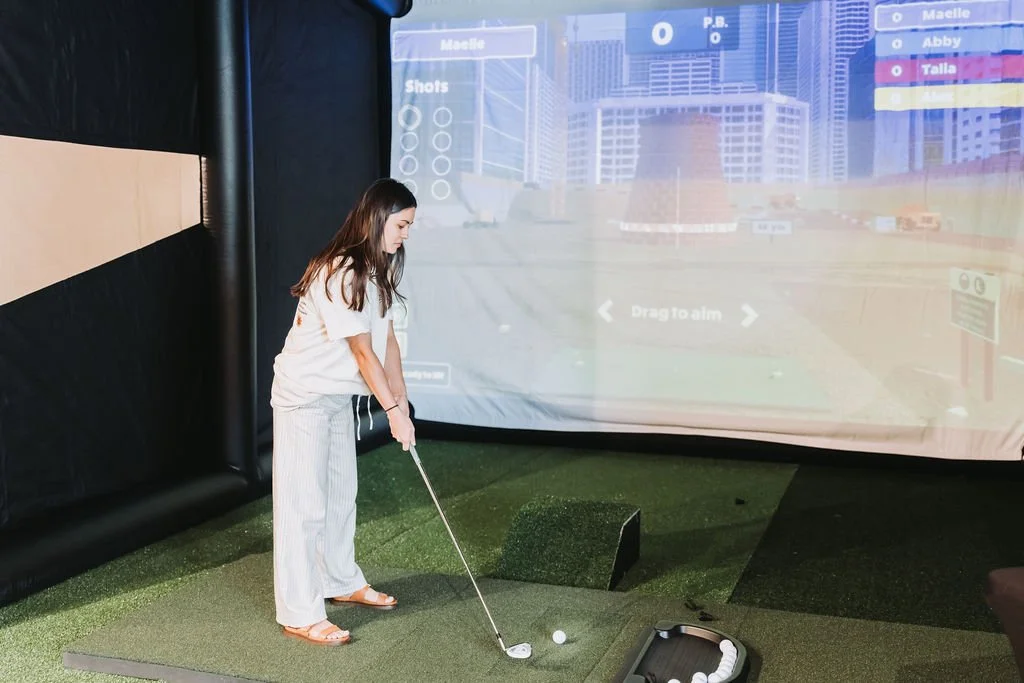 Woman practicing golf in indoor simulator, standing on a green mat, holding a golf club, with a projected golf course on the screen behind her.