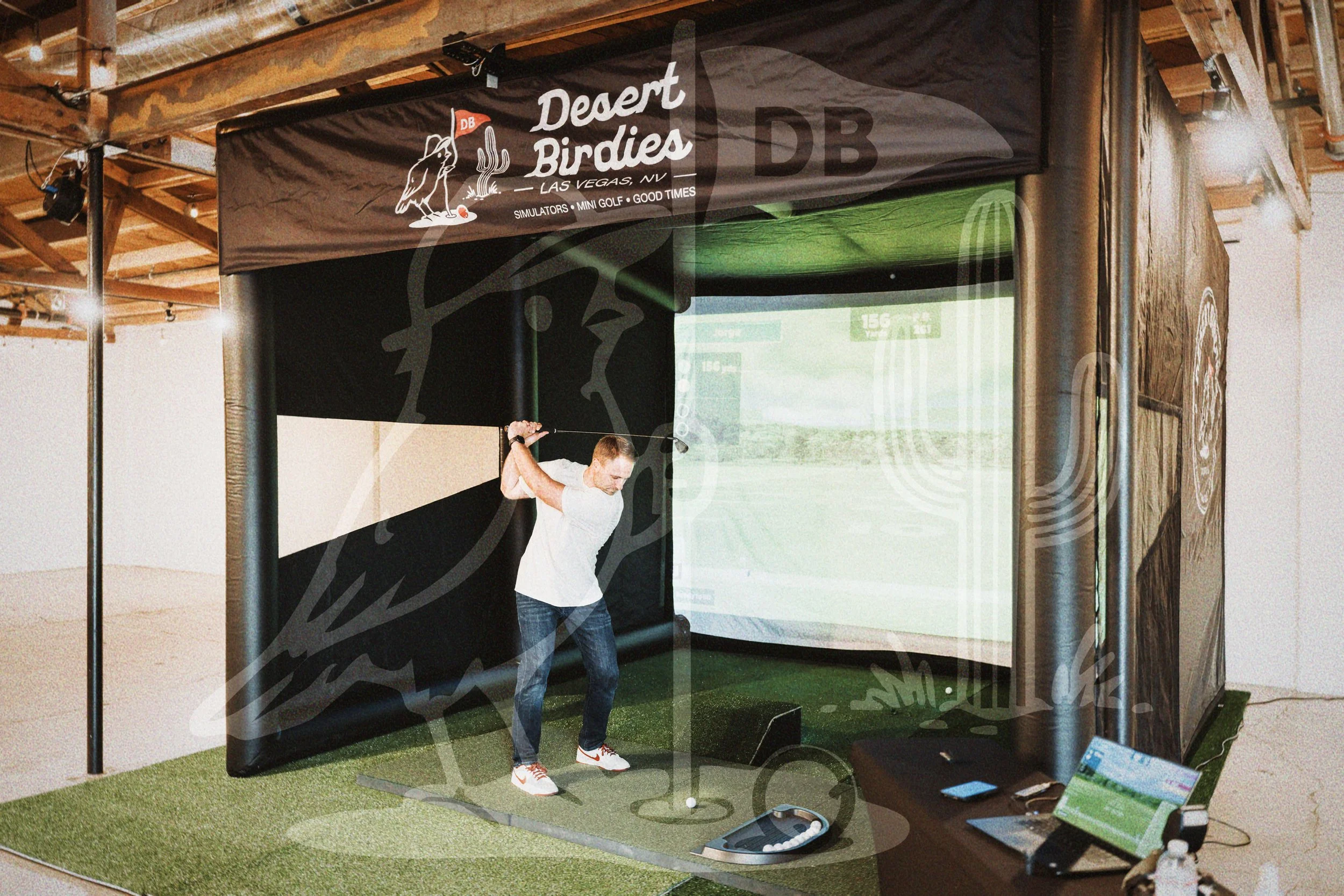 Man playing virtual golf game at indoor simulation center, brand called Desert Birdies, with screen showing golf course, golf balls on floor, and computer setup nearby.