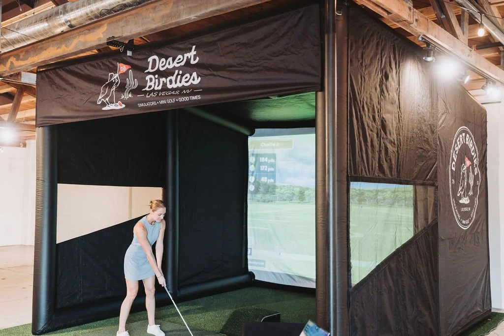 An indoor golf simulator room with a woman playing golf. The room has black padded walls with a golf-themed sign reading 'Desert Birds Las Vegas NV.' The simulator screen shows a golf course, and the room features a turf mat and a ceiling with exposed wooden beams.