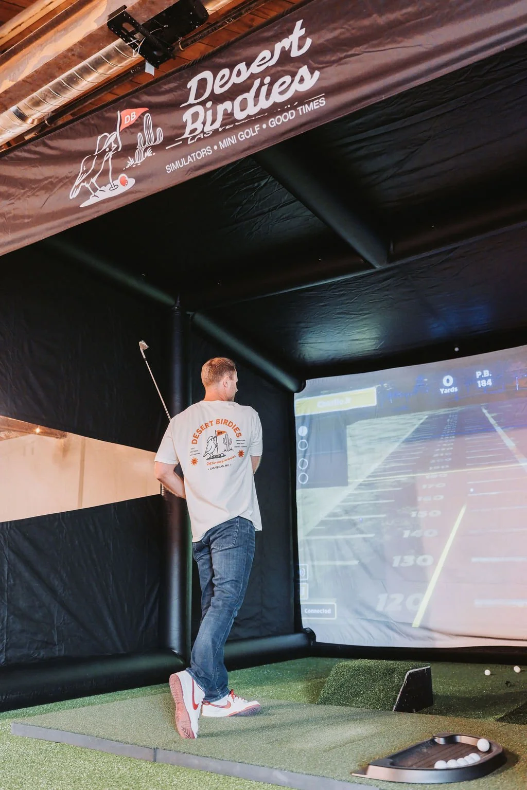 An indoor golf simulator at Desert Birdies, with a man preparing to take a shot, wearing a Desert Birdies t-shirt. The simulator screen displays a golf course.
