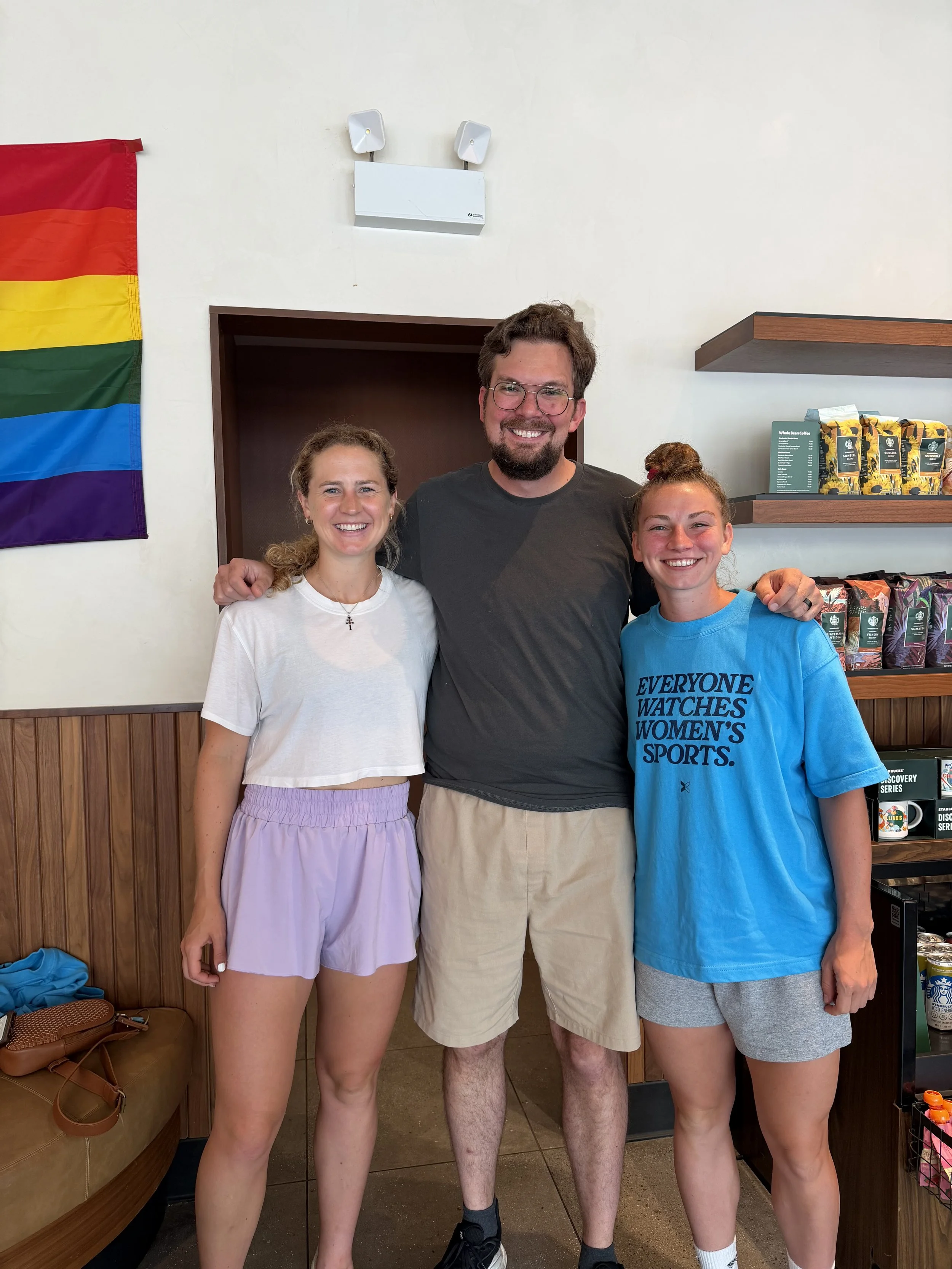 Group of three smiling young adults standing indoors near a wooden wall and a shelf with coffee bags, with a rainbow pride flag hanging on the left side.
