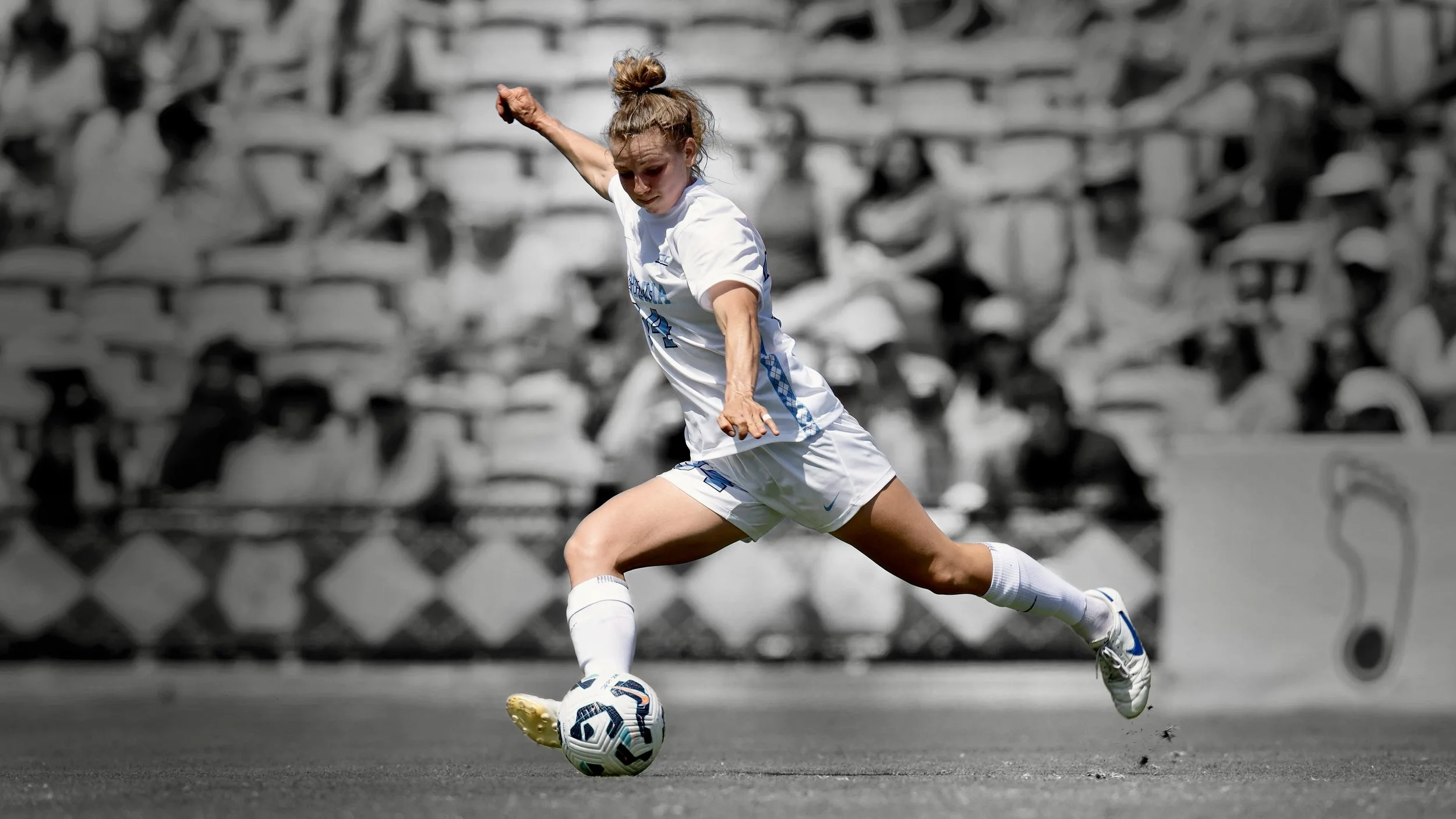 Female soccer player in white uniform kicking a soccer ball on the field with an empty stadium in the background.