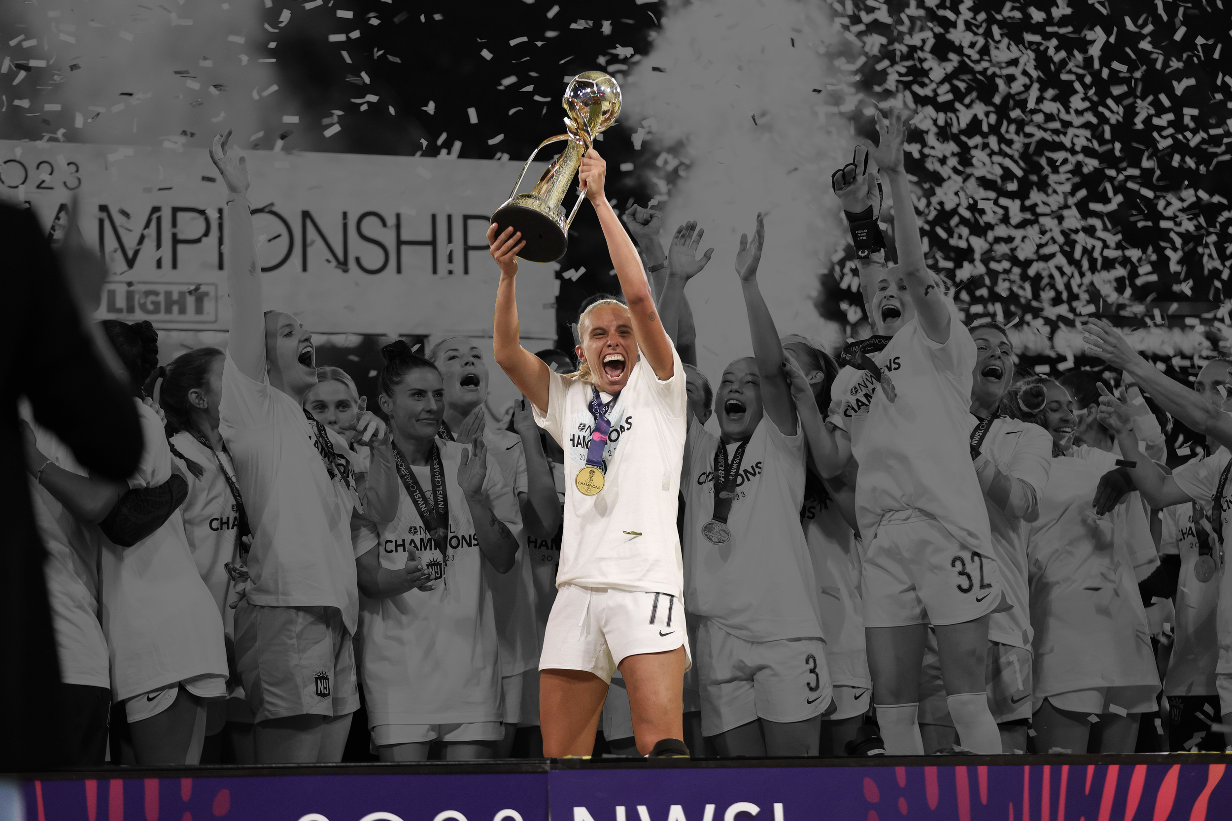 A sports team celebrating a victory with some members holding medals and a trophy, and a woman in the center holding up the trophy, surrounded by cheering teammates and confetti.