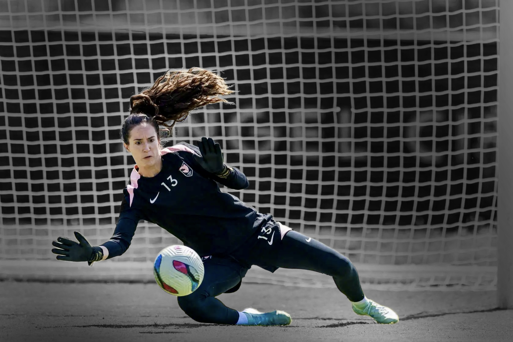  Female soccer goalkeeper in black uniform diving to her left to block a colorful soccer ball in front of a goal net.
