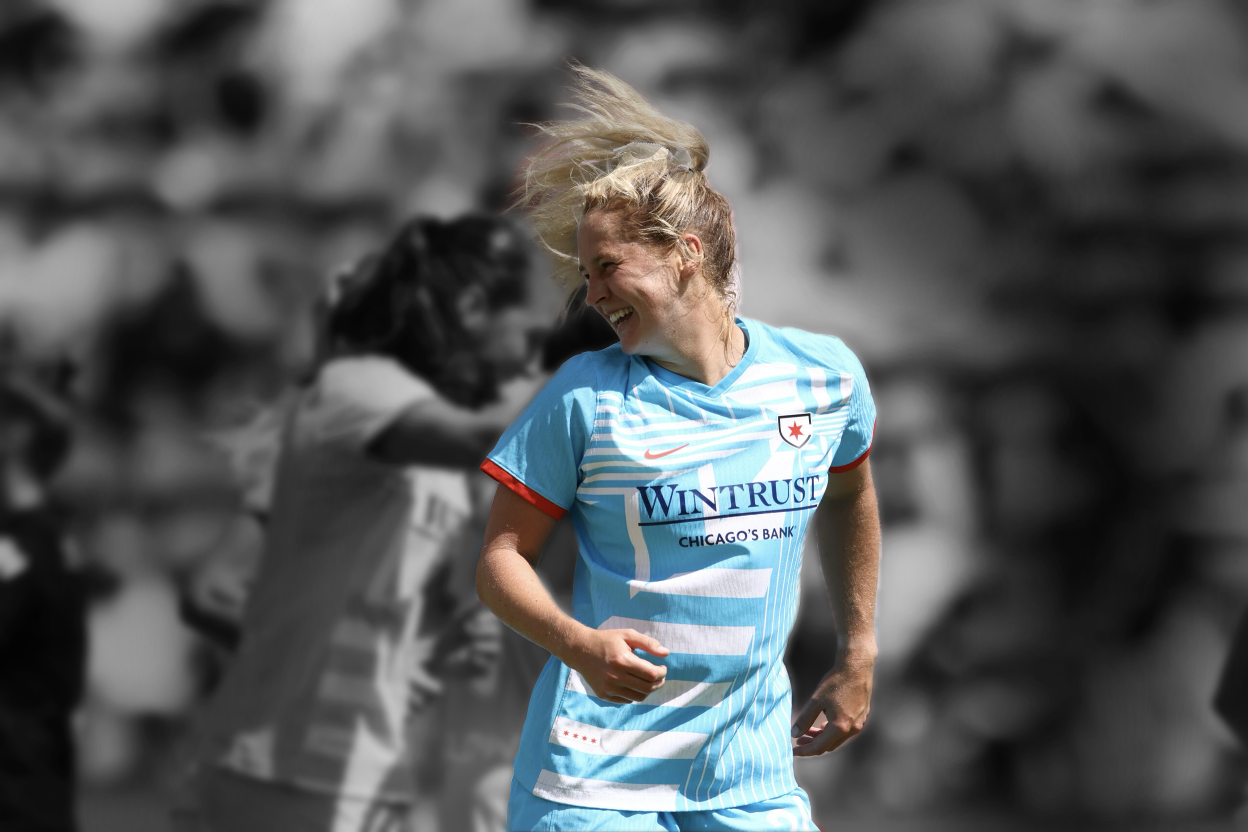 A female soccer player with blonde hair wearing a light blue uniform smiling and running on the field, with a blurred background of other players.