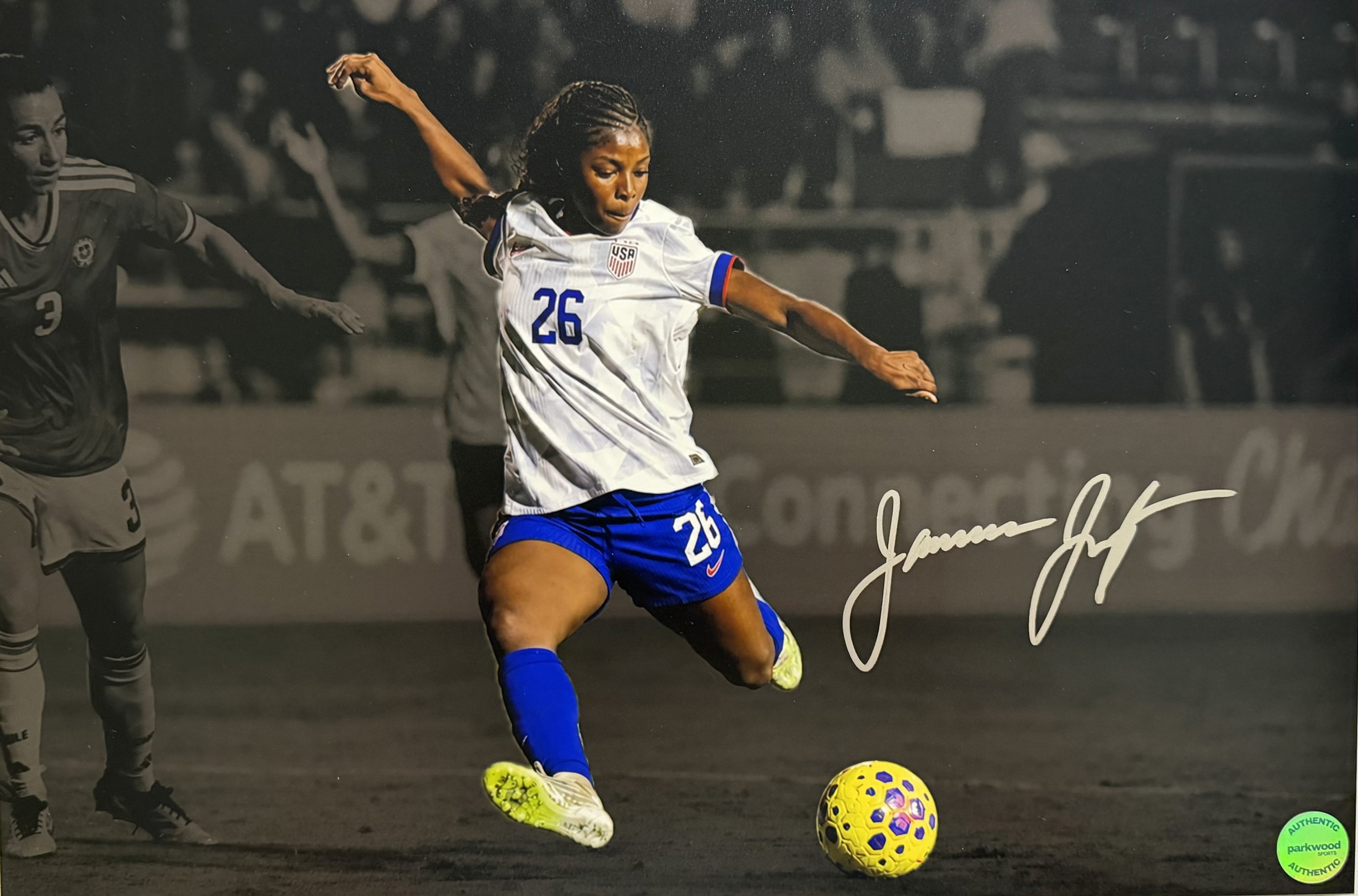 A female soccer player in a white and blue uniform with the number 26 is kicking a yellow and purple soccer ball during a match on the field, with another player in a dark uniform nearby and spectators in the background.