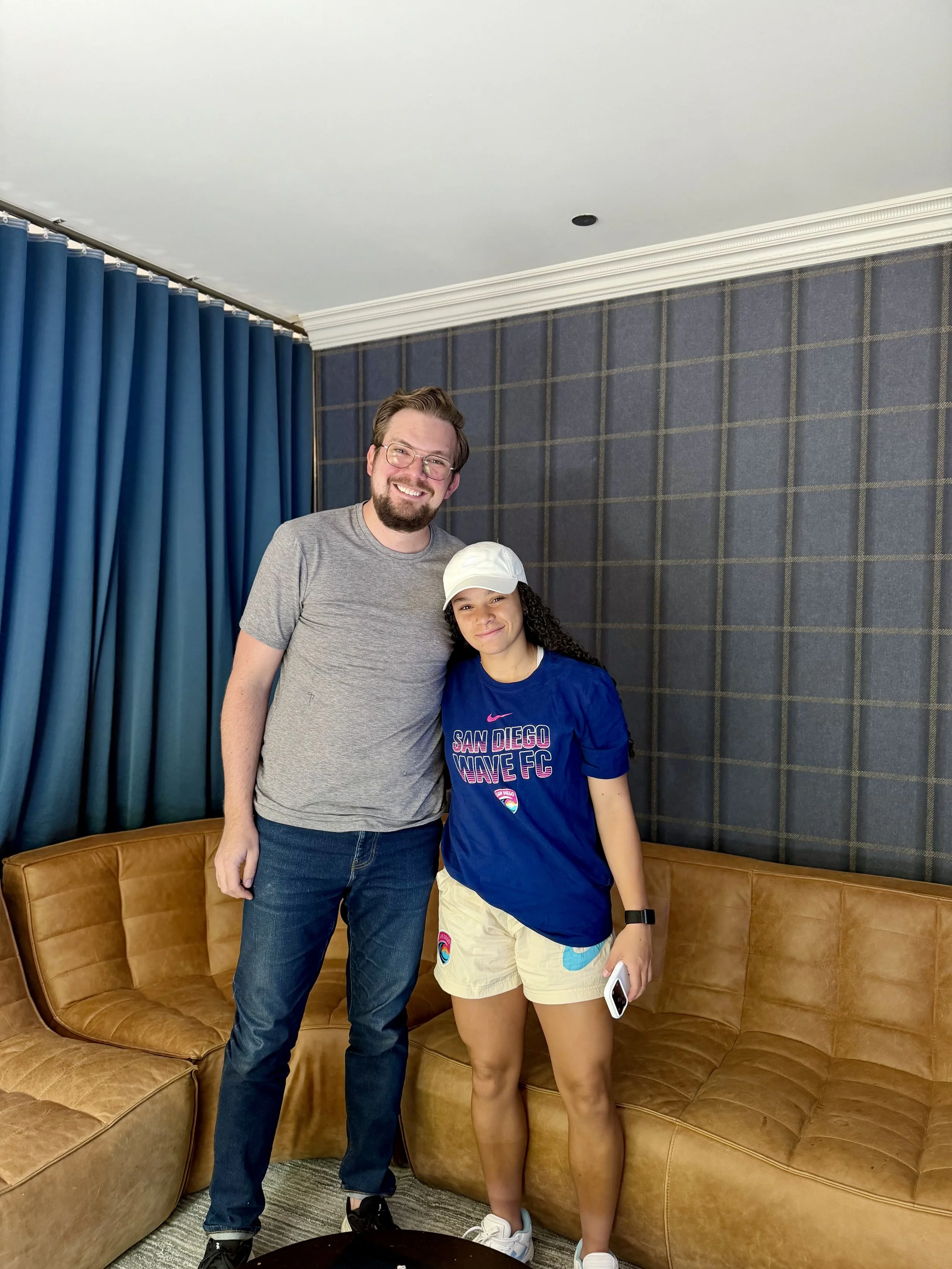 A man and a woman with a tattooed arm standing together on a tan leather couch in a room with blue curtains and dark plaid wallpaper, smiling at the camera.