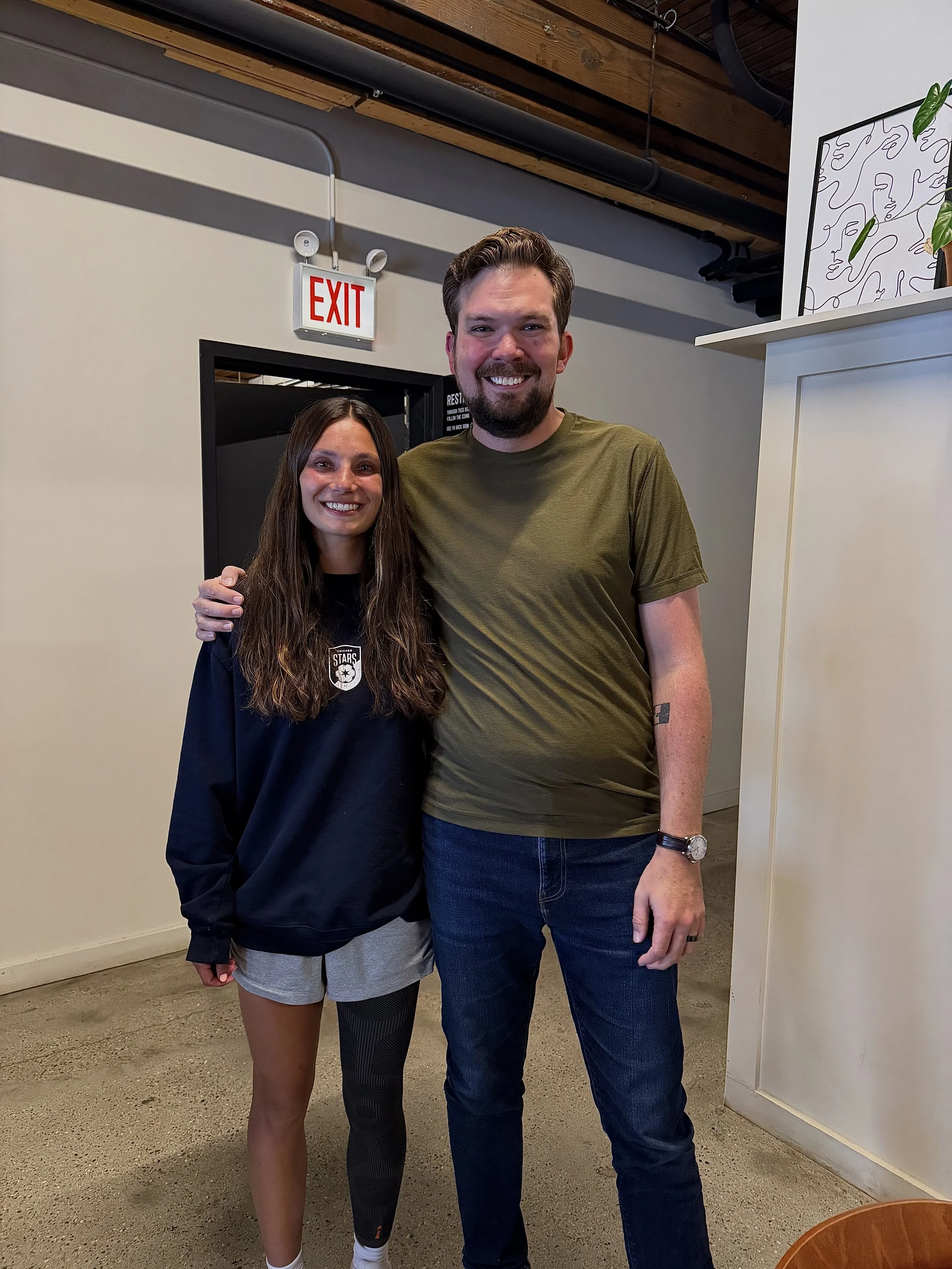 A man and woman standing indoors, smiling for a photo. The woman has long dark hair, is wearing a black shirt with a logo, and gray shorts. The man has short brown hair, a beard, and is wearing a green t-shirt and jeans. They are standing close to each other, with the woman’s arm around the man's back.