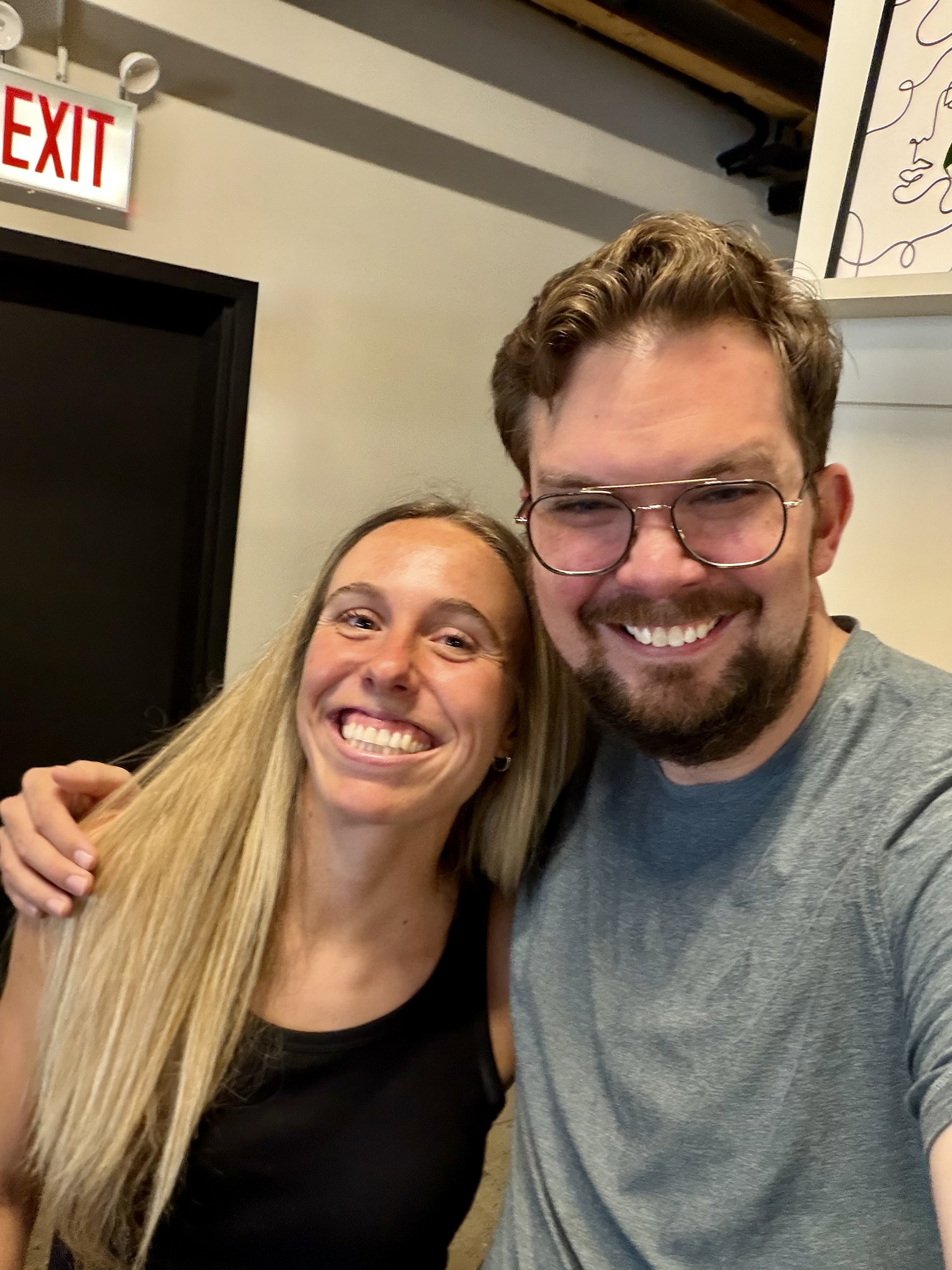 A smiling woman with long blonde hair and a man with short curly hair, glasses, and a beard, taking a selfie together in an indoor setting.