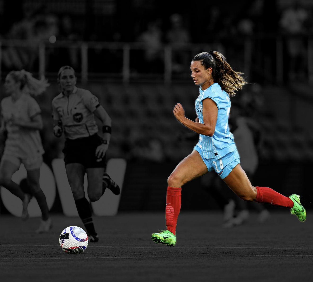 A female soccer player in a light blue uniform with red socks and green cleats is running on the field, with a soccer ball in front of her. The background of the image is black and white, with two women and a referee visible in the distance.