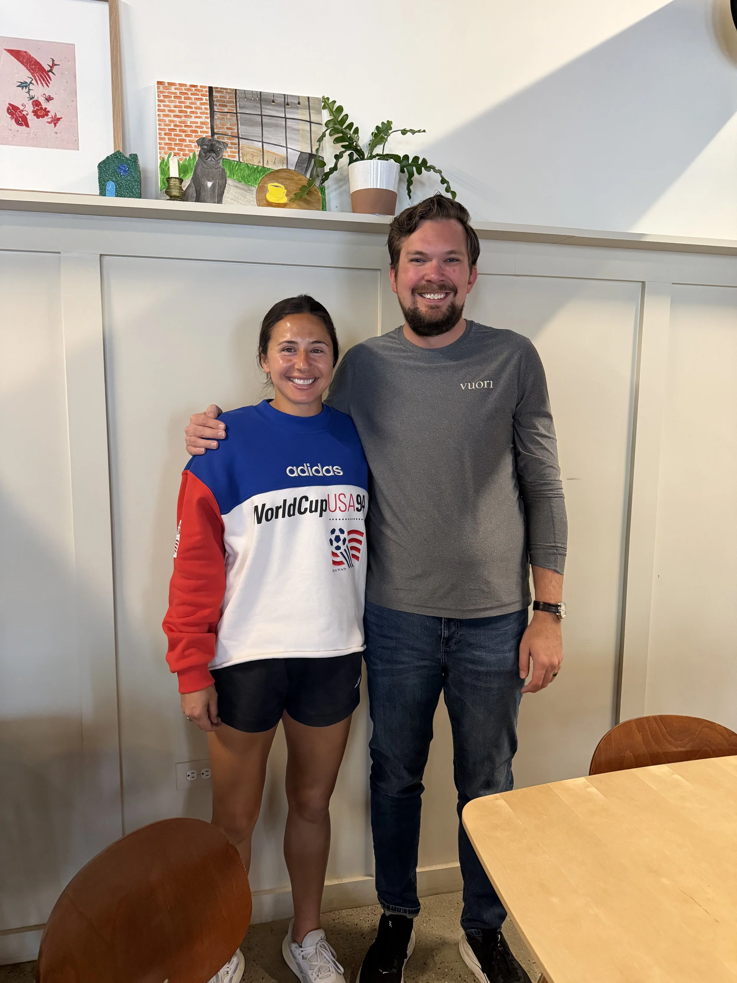 A woman and a man are standing close together indoors, smiling at the camera. The woman is wearing a blue, white, and red Adidas sweatshirt and black shorts, while the man is dressed in a gray long-sleeve shirt and dark jeans. There are chairs and a table in the room, and artwork and potted plants are visible on a shelf behind them.