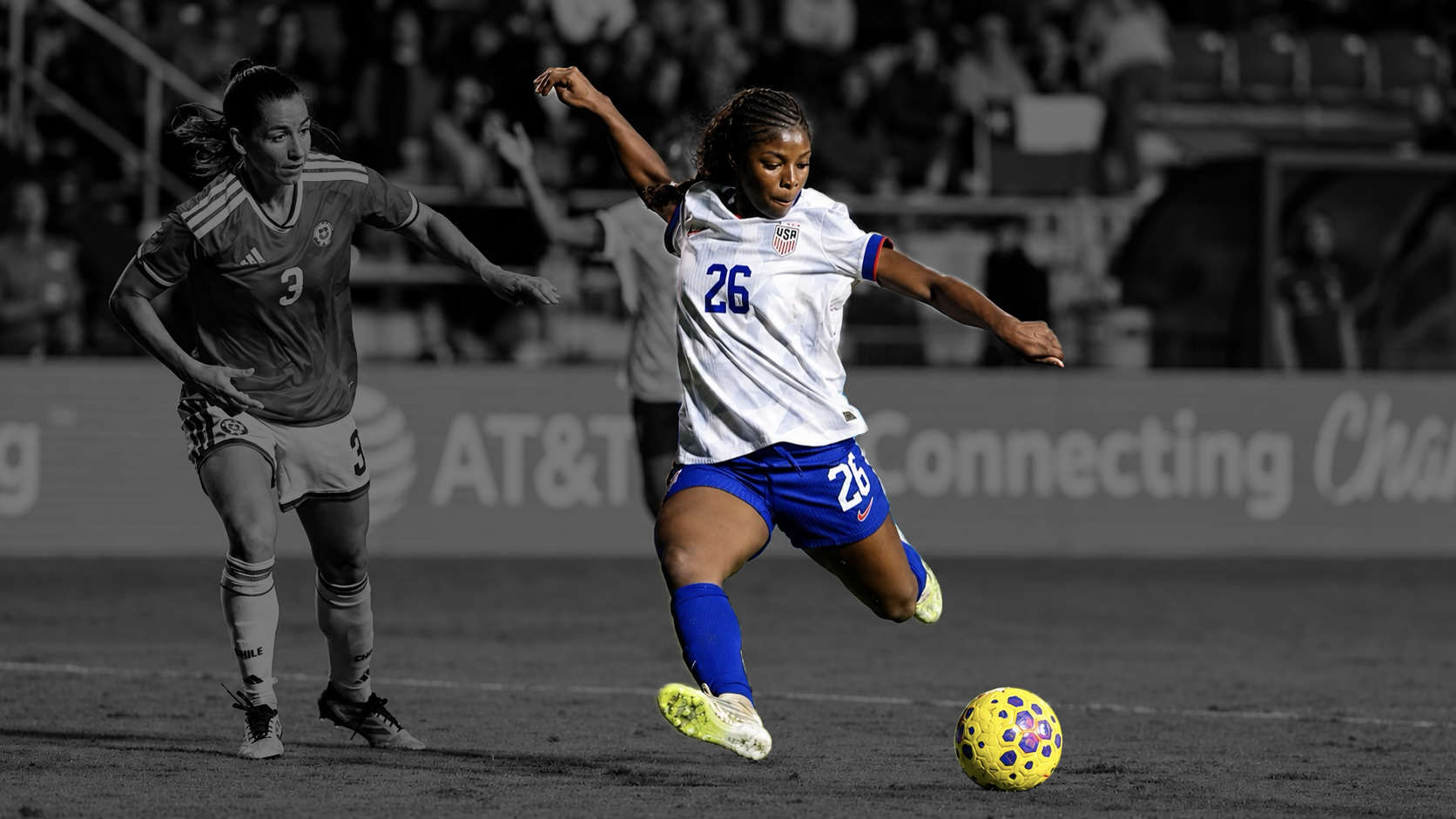 A female soccer player in a white and blue uniform with the number 26 is kicking a yellow and purple soccer ball during a match on the field, with another player in a dark uniform nearby and spectators in the background.