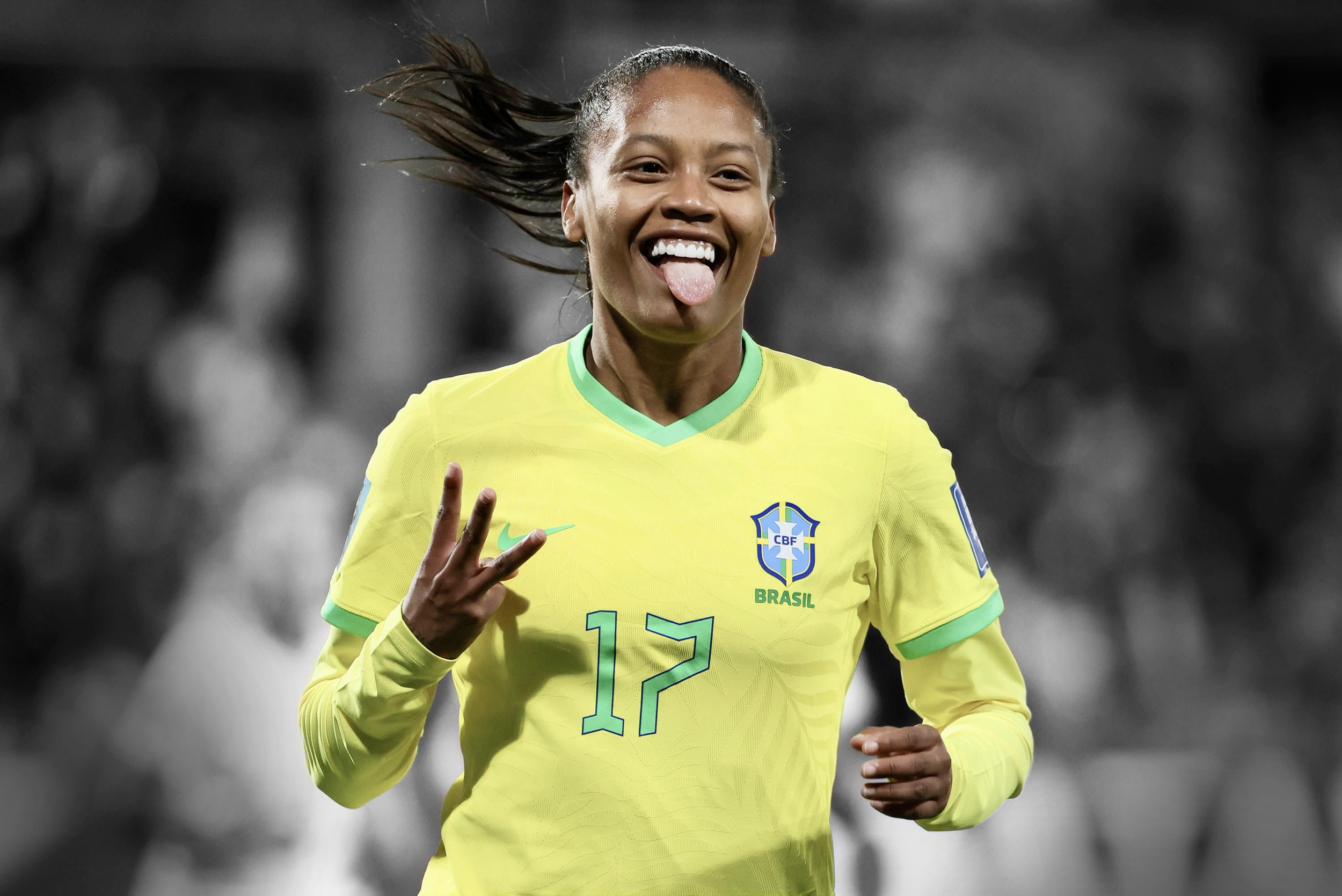 A woman wearing a yellow Brazilian soccer jersey with green accents, smiling, sticking her tongue out, and making a peace sign gesture with her left hand during a soccer match.