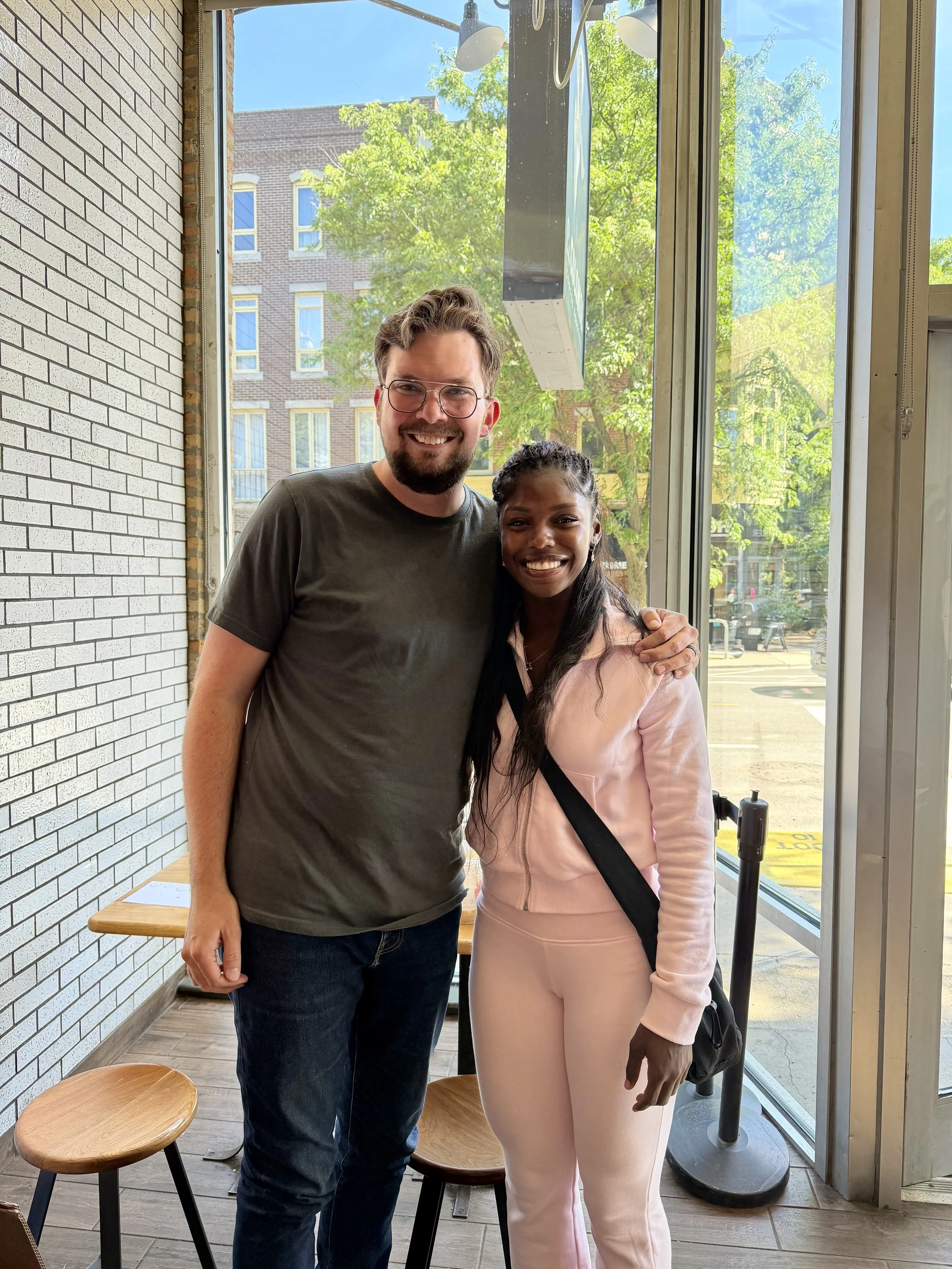 A man and woman standing inside a cafe, smiling, with a large window behind them showing trees and a brick building outside.