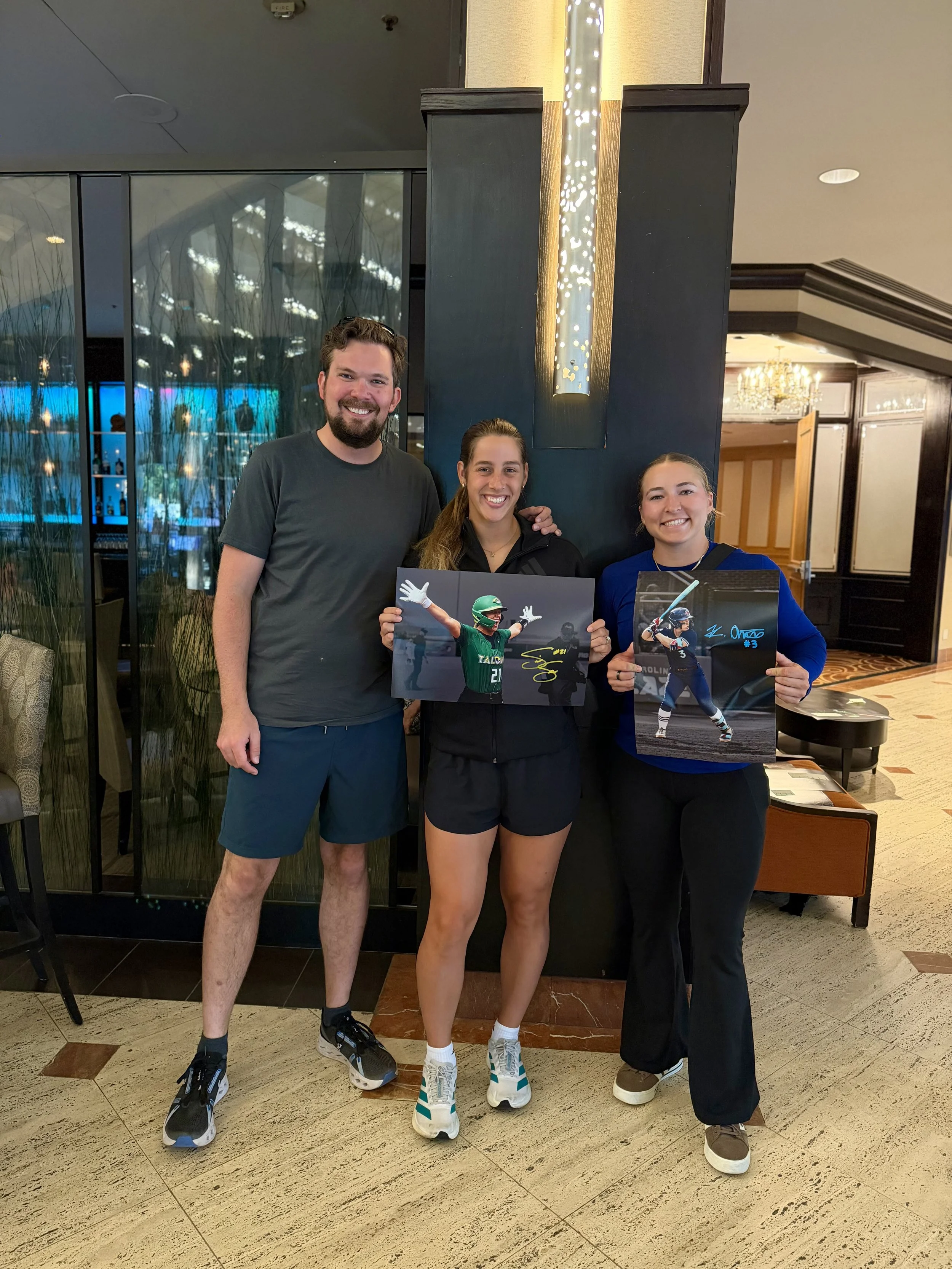 Three people standing indoors, smiling at the camera. The person on the left is a man with dark hair and a beard, wearing a gray t-shirt and dark shorts. The middle person is a woman with long brown hair, wearing black athletic clothing, holding a signed photo of a football player. The person on the right is a woman with blonde hair, wearing a blue shirt, holding a signed photo of a female athlete. They are standing in front of a dark wall with decorative lighting, with a chandelier visible in the background.