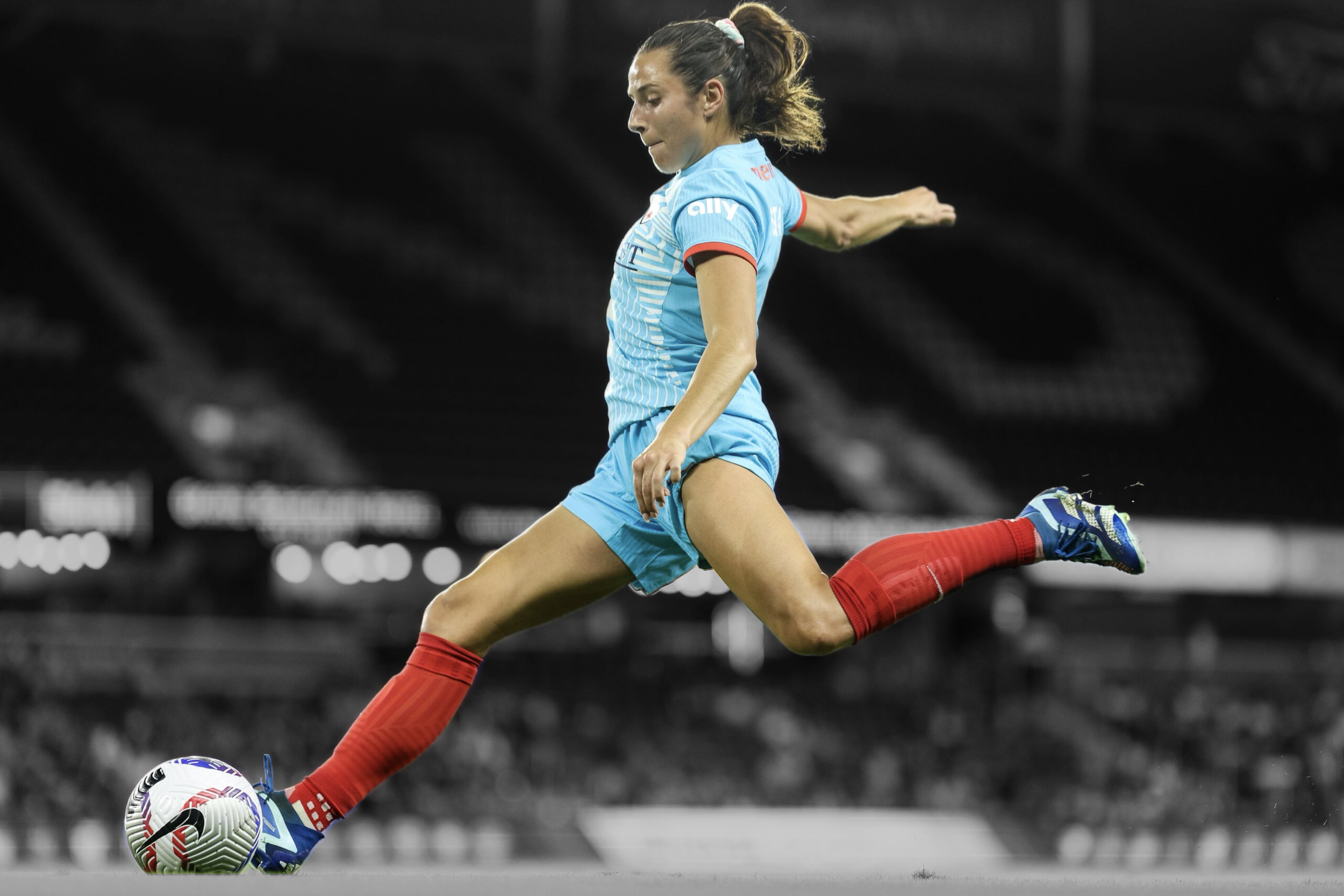 A female soccer player in a blue uniform, red socks, and blue shoes is kicking a soccer ball on a soccer field, with a dark and blurred stadium background.