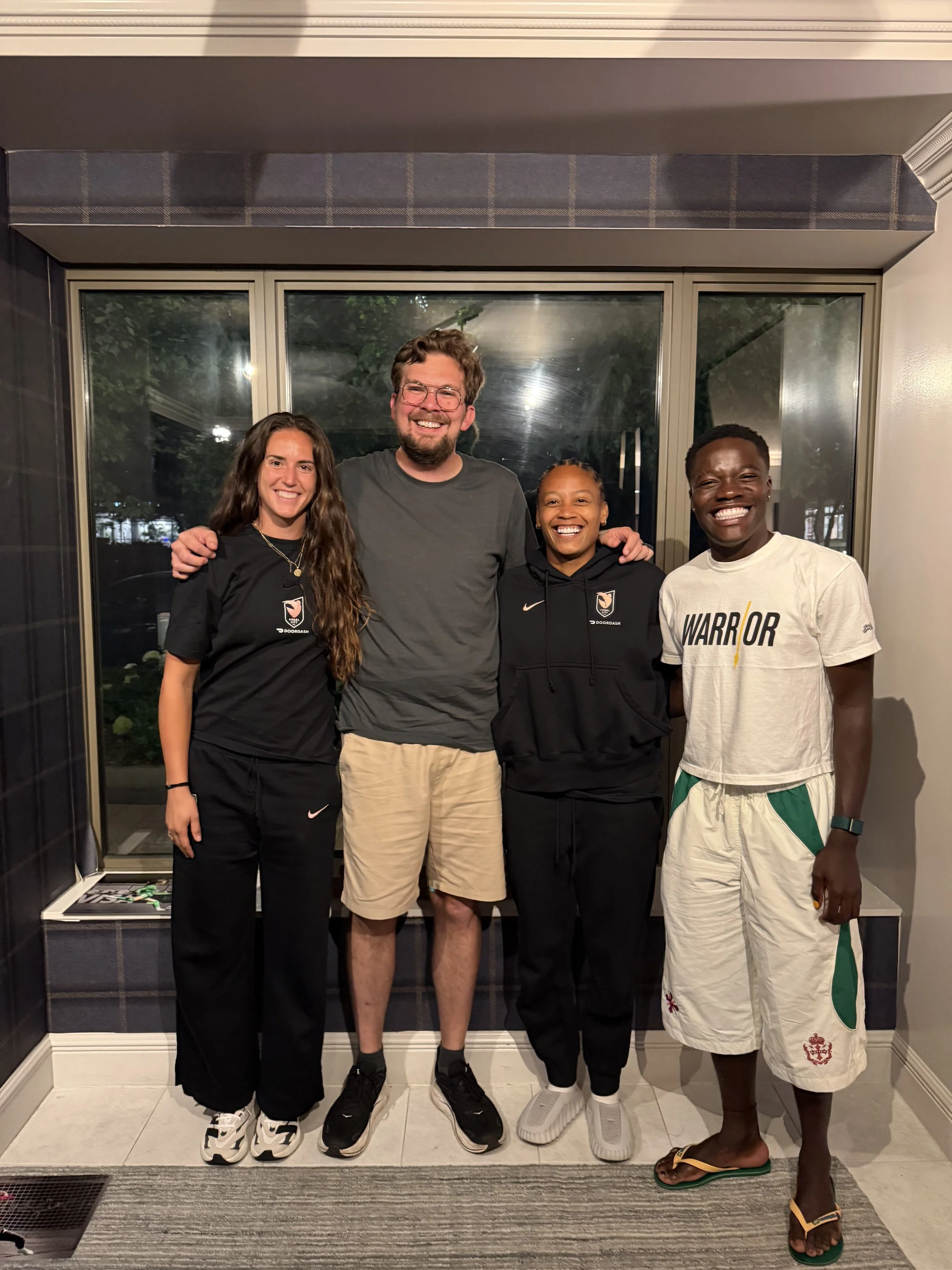 Four young people standing together in a row, smiling and posing for a photo indoors in front of large windows with a night scene outside. They are dressed casually, with some wearing athletic or team apparel.