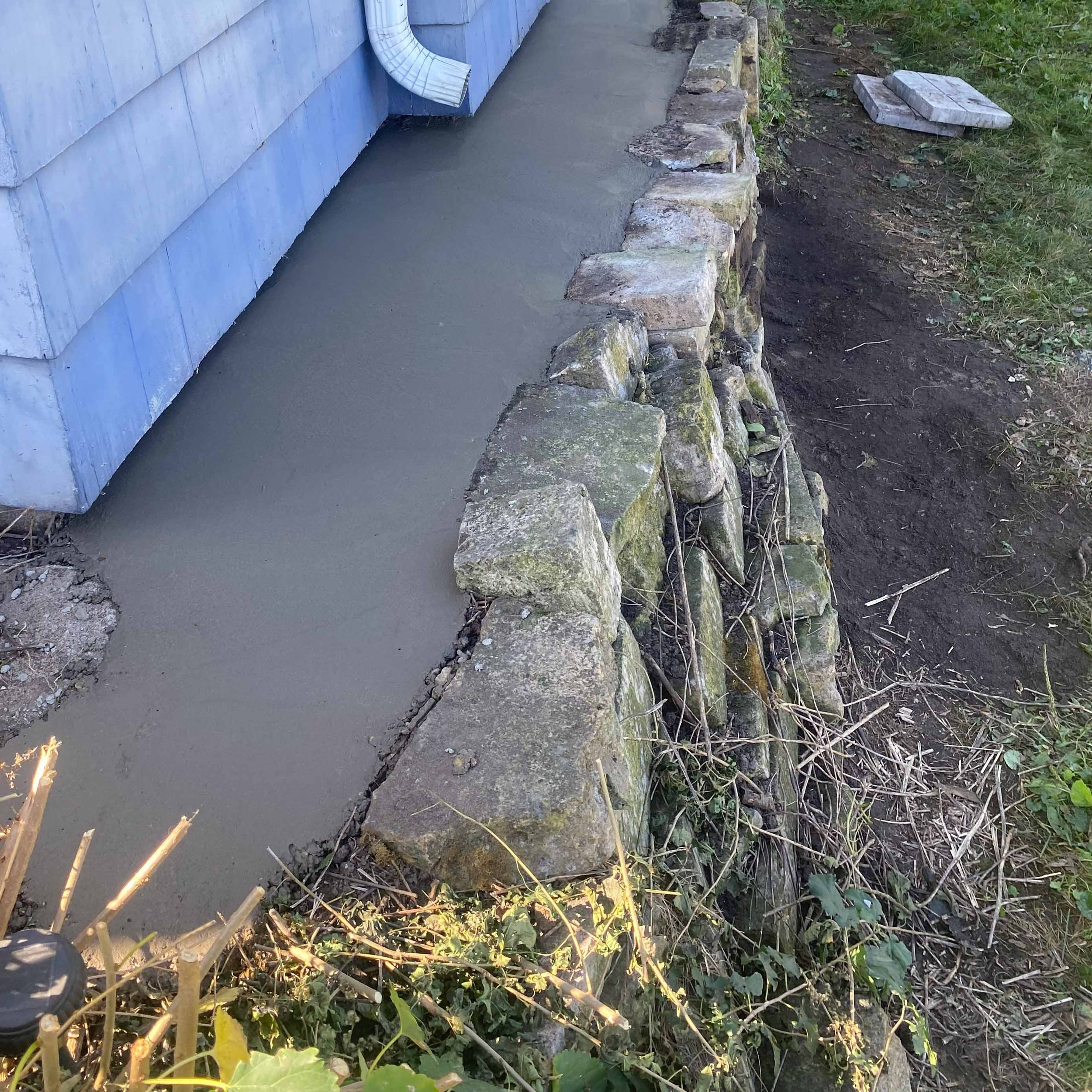 Concrete pathway next to a house foundation with stone border, grassy area with plants, and a partially visible drainage pipe.
