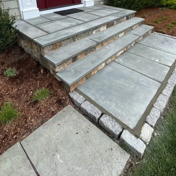 Concrete steps leading up to a porch with large stone pavers and a stone walkway extending to the yard, bordered by small bushes and mulch.
